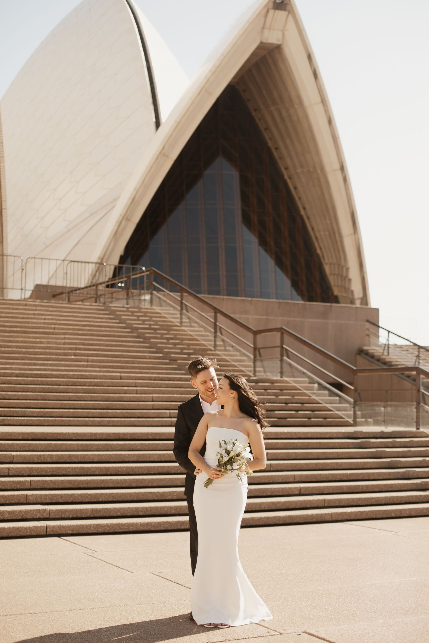 Wedding couple poses on steps of iconic Sydney Opera House in elegant attire.
