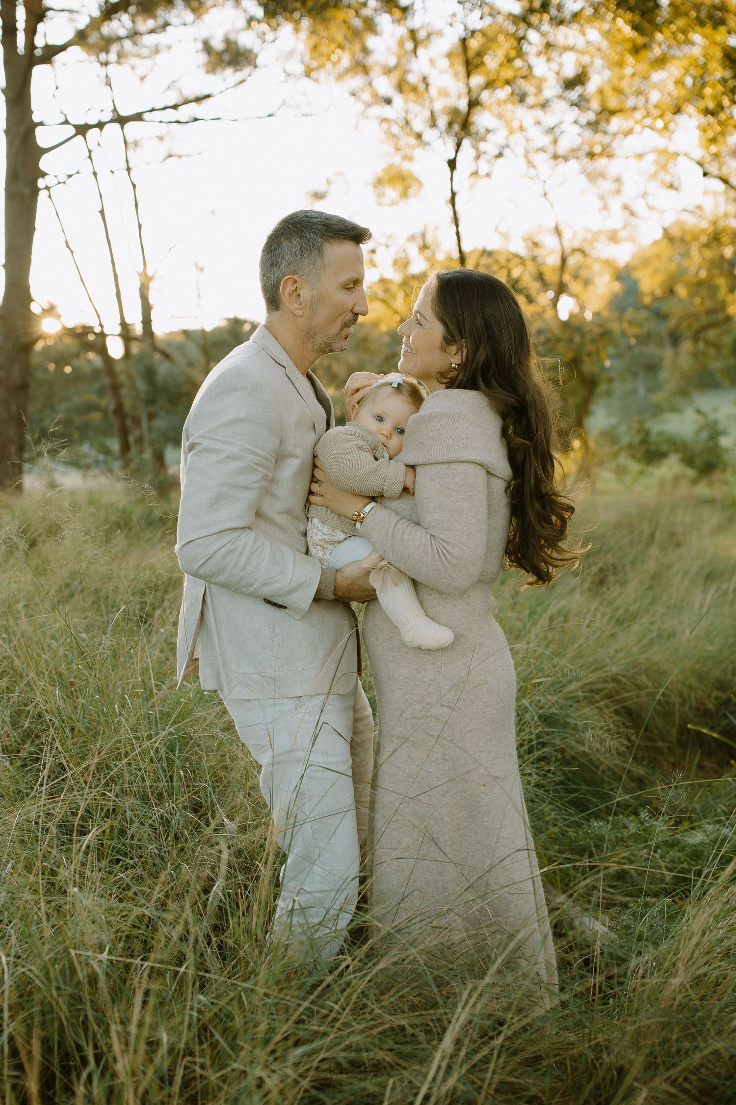 A couple in neutral outfits embraces while holding their baby during a sunset photoshoot in tall grass.