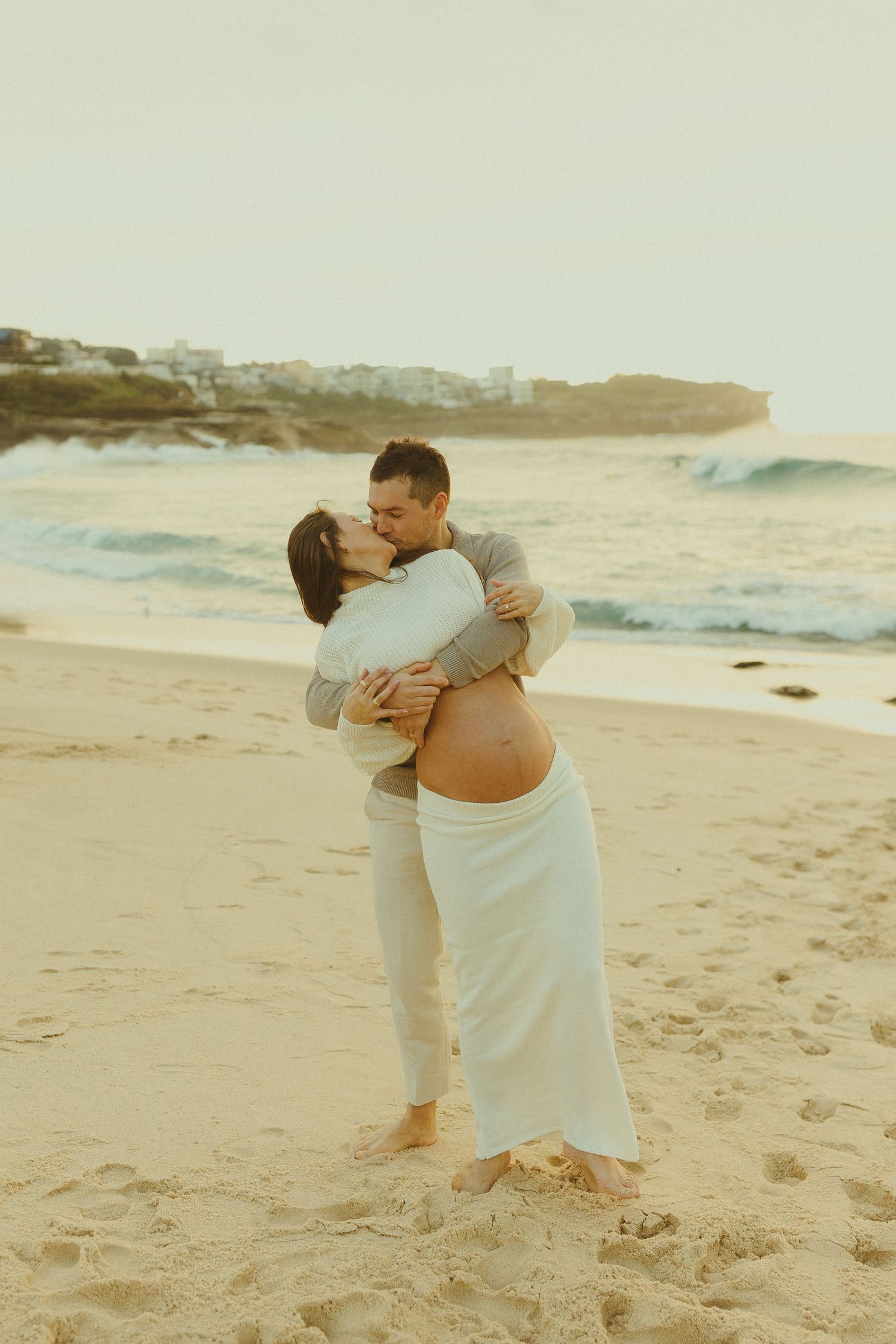 A romantic moment captured between couple embracing on a sandy beach at sunset with ocean waves in the background.