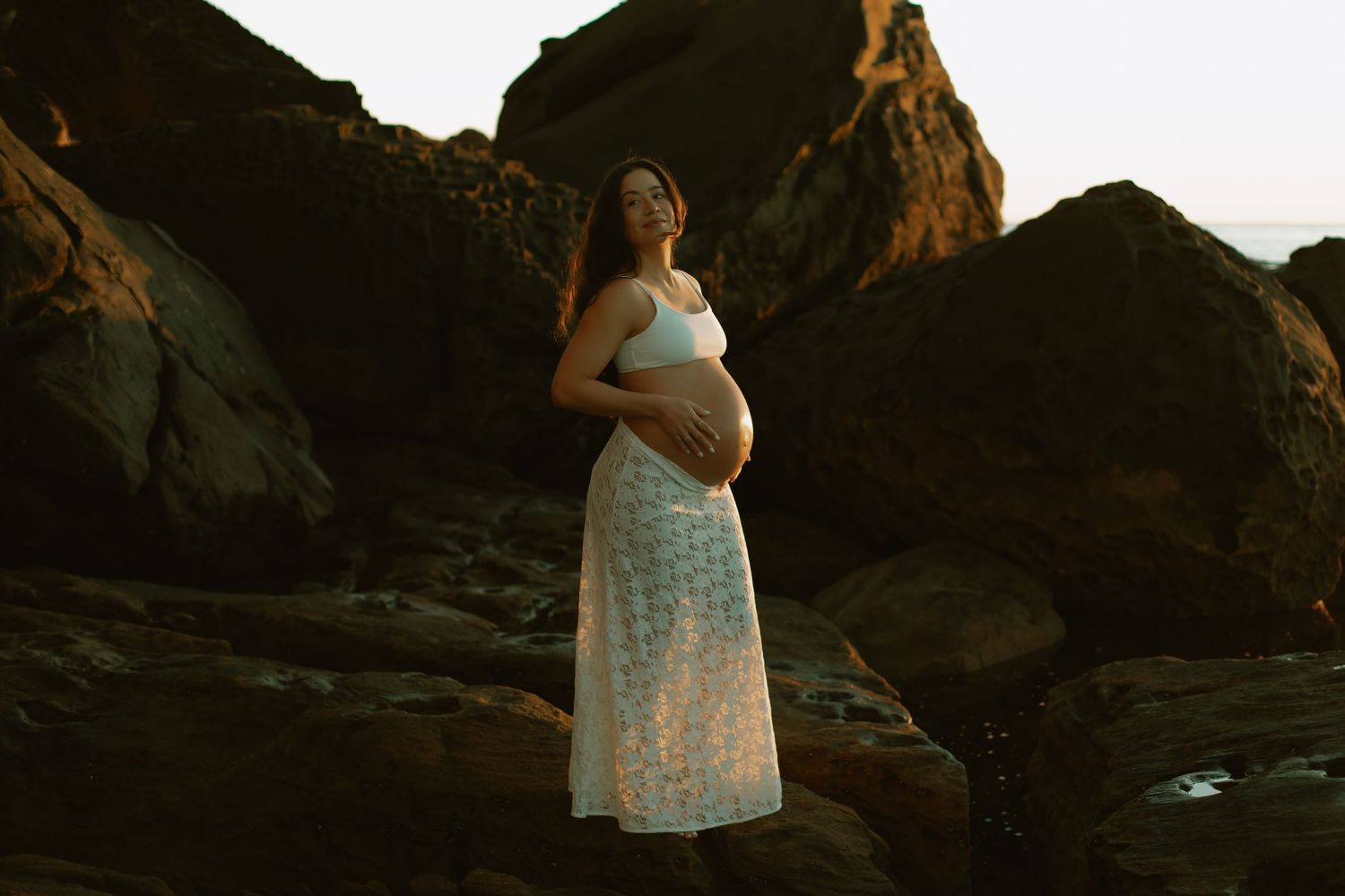 Expectant woman in lace dress poses against dramatic rocky landscape during sunset photoshoot.