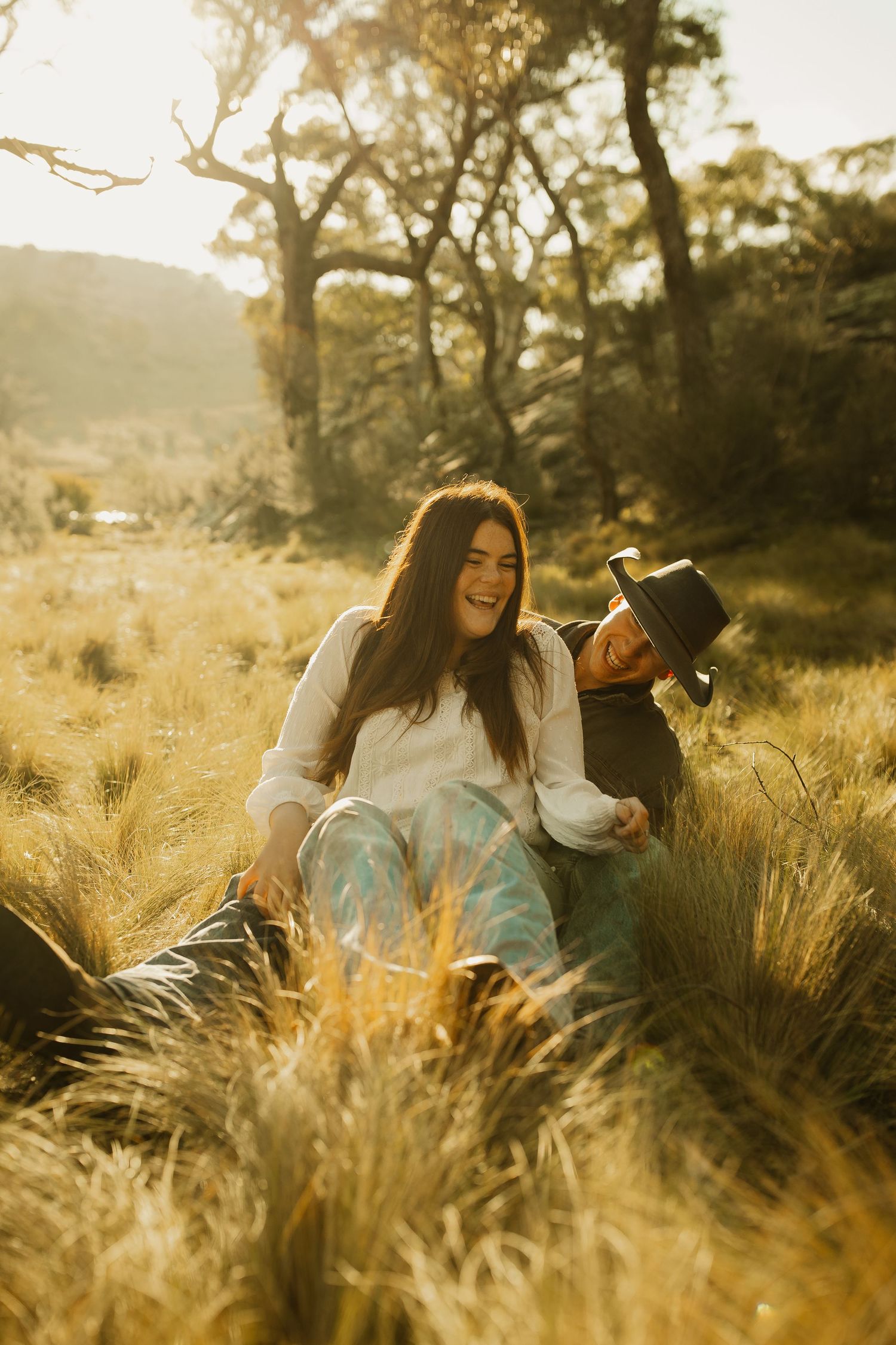 A couple shares a joyful moment while sitting in tall golden grass during sunset.