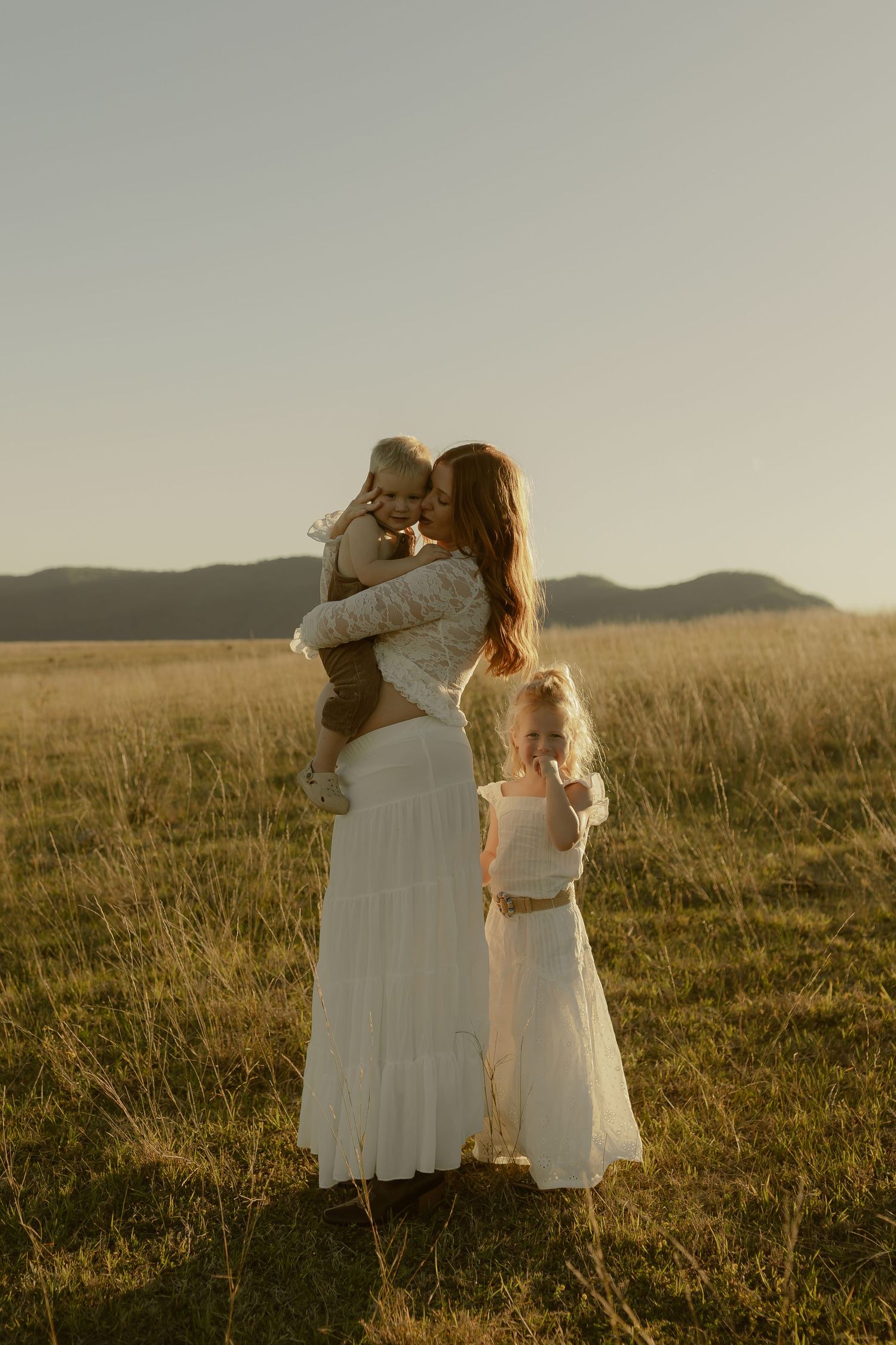 A mother in a white dress holds her baby while her young daughter stands beside her in a golden field at sunset.