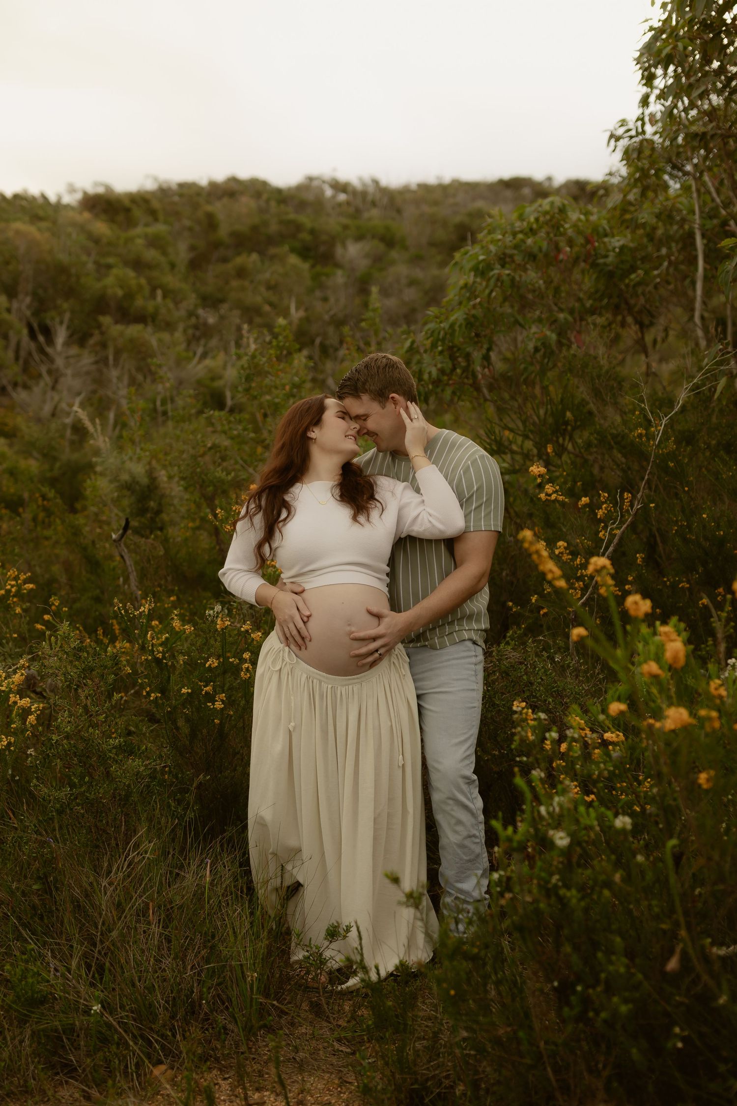 Couple sharing a tender moment in lush greenery, woman in cream dress showing baby bump, maternity photo shoot outdoors.