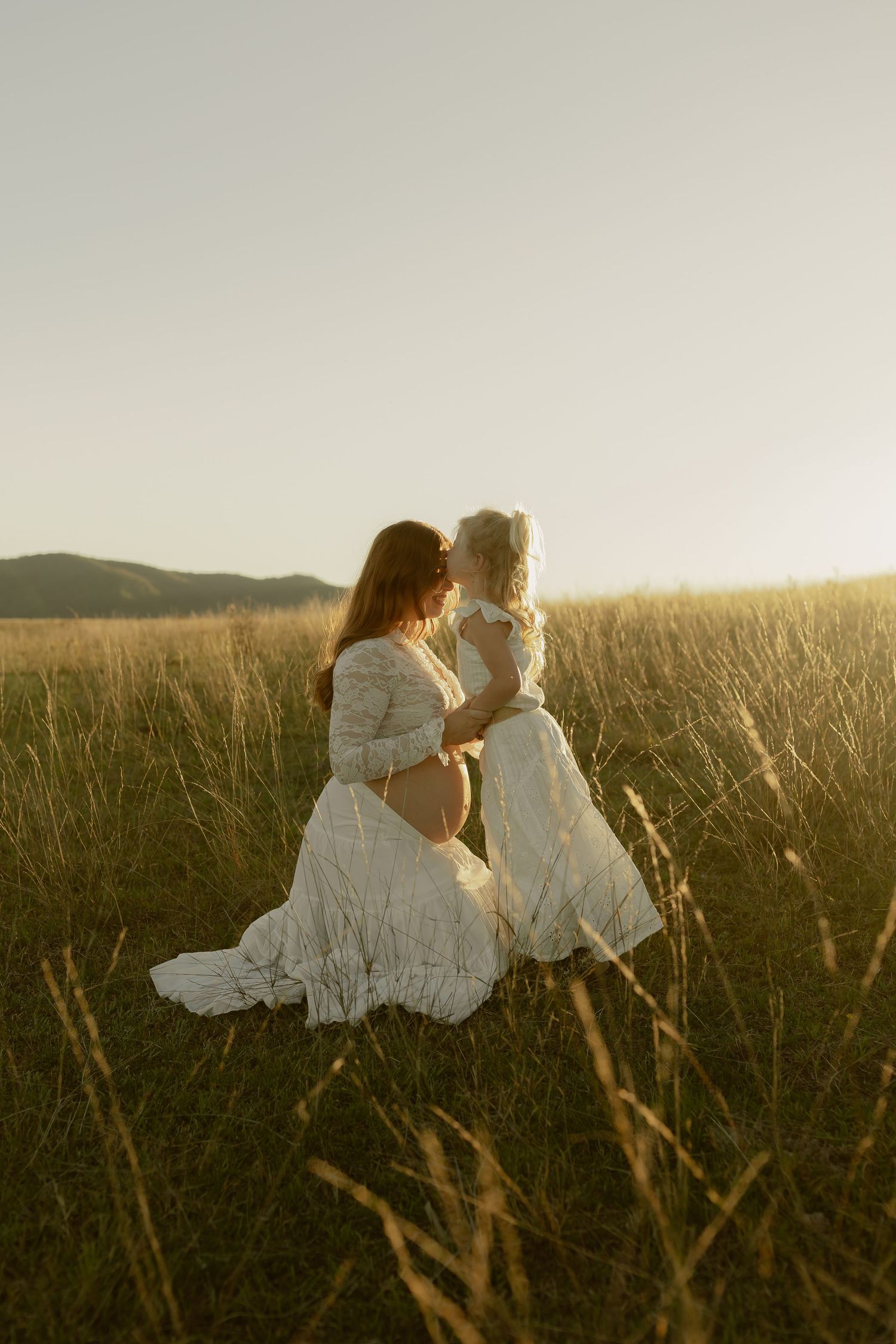Pregnant woman and toddler in matching white lace dresses share a tender moment in a golden sunlit field.