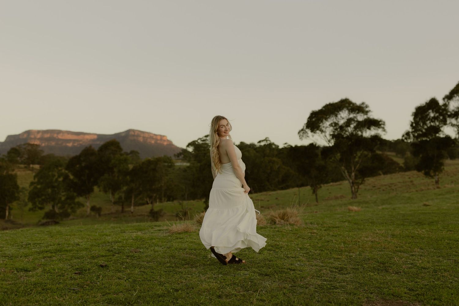 Bride in flowing white dress twirling on green hillside at dusk with mountain backdrop in rural Australia.