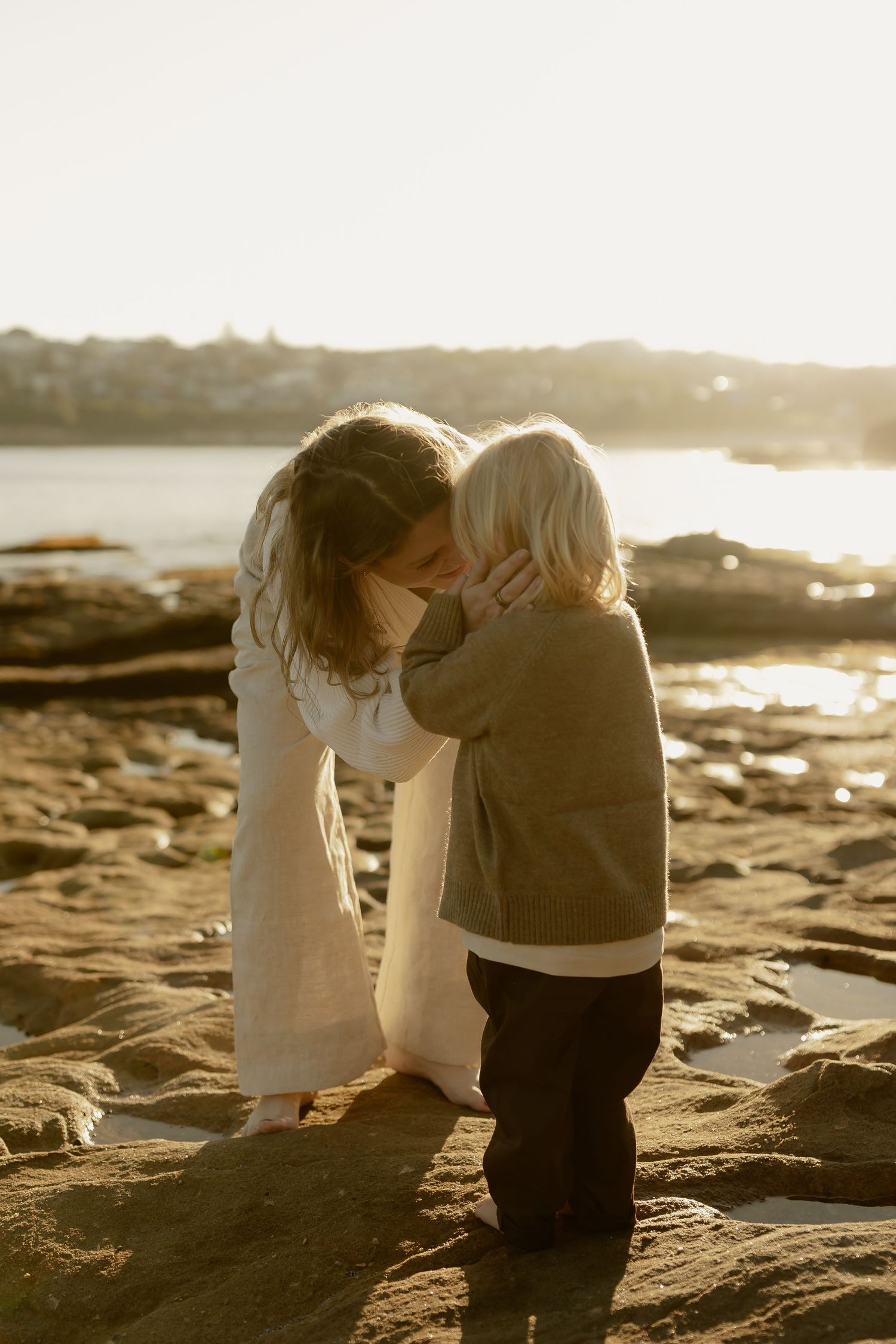 Mother and young child sharing a tender forehead kiss on a rocky beach at golden sunset.