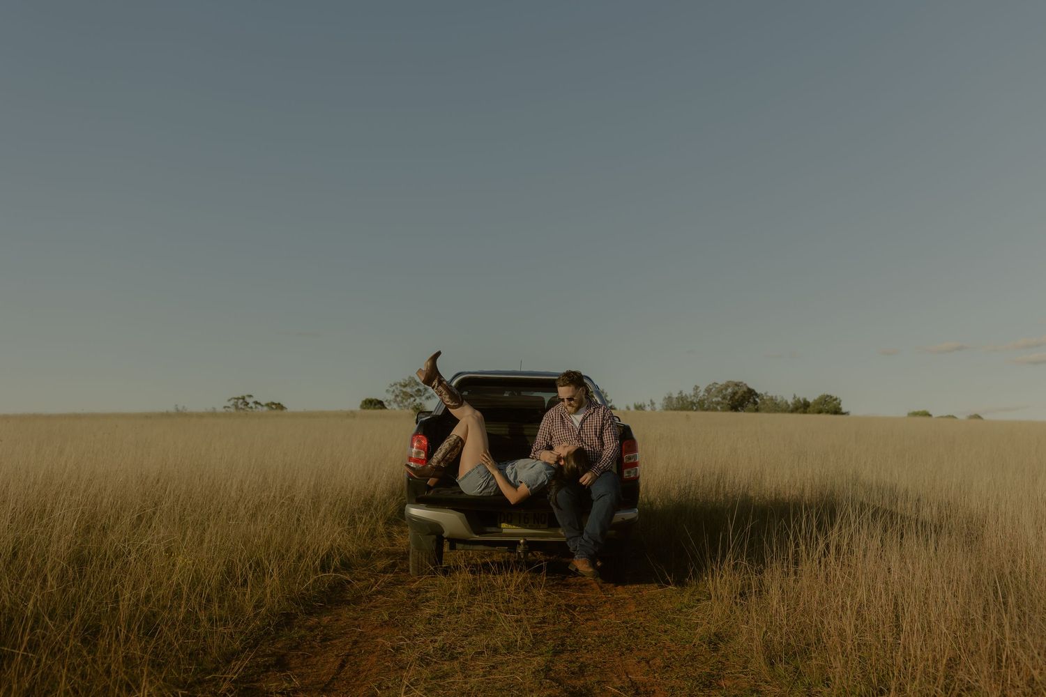 Two people relaxing on a pickup truck tailgate surrounded by golden grass fields under a clear blue sky.