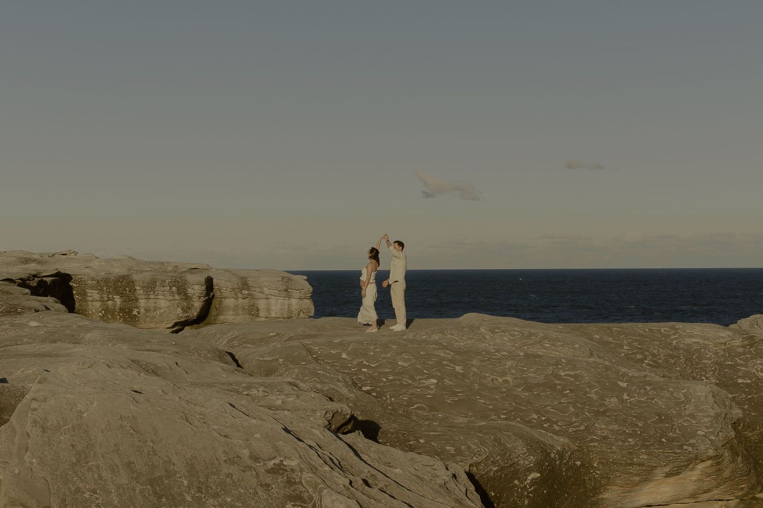 Couple holding hands on rocky coastal cliffs with ocean horizon at golden hour.