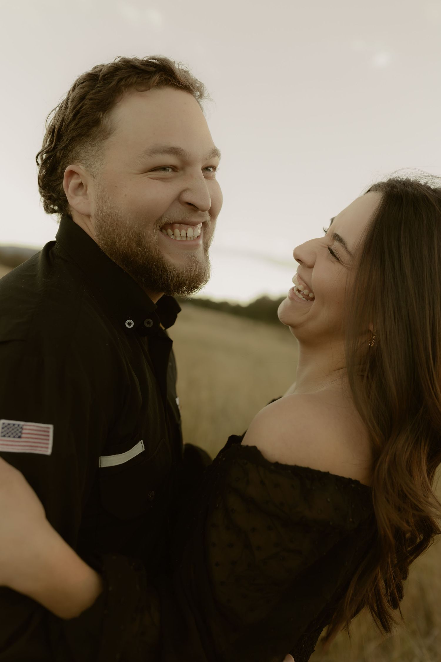 Happy couple embracing and laughing together outdoors in a golden field at sunset, wearing matching black outfits.