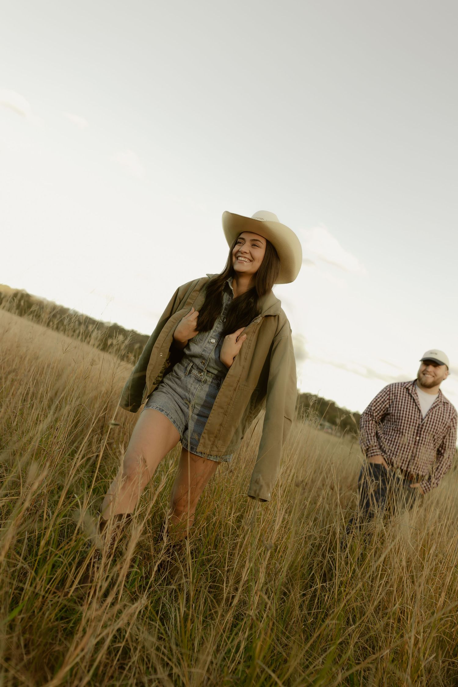 Woman in cowboy hat and denim shorts walks through golden field with man in plaid shirt at sunset.