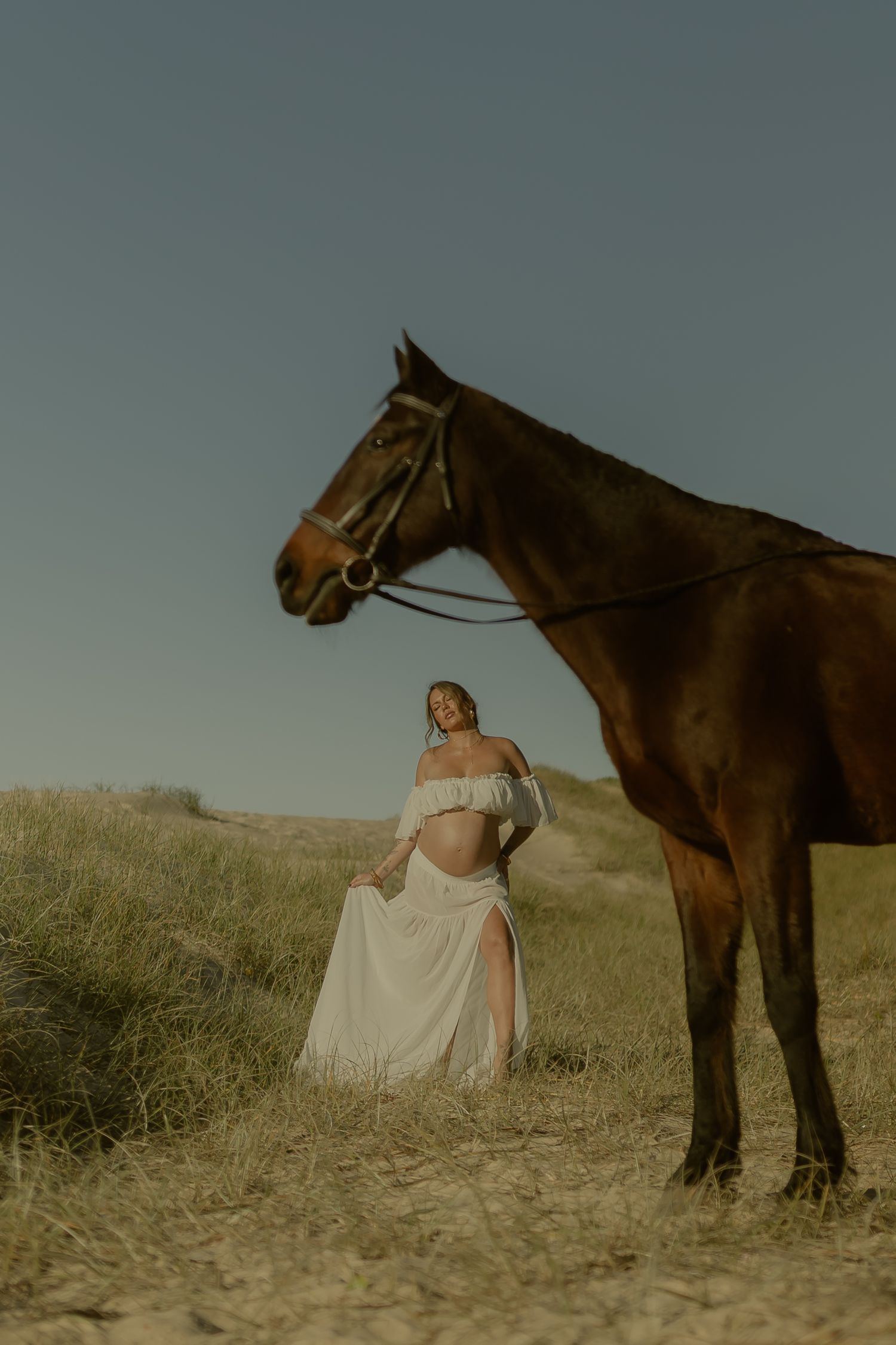 Pregnant woman in white off-shoulder gown poses beside a brown horse in a grassy field under a clear sky.