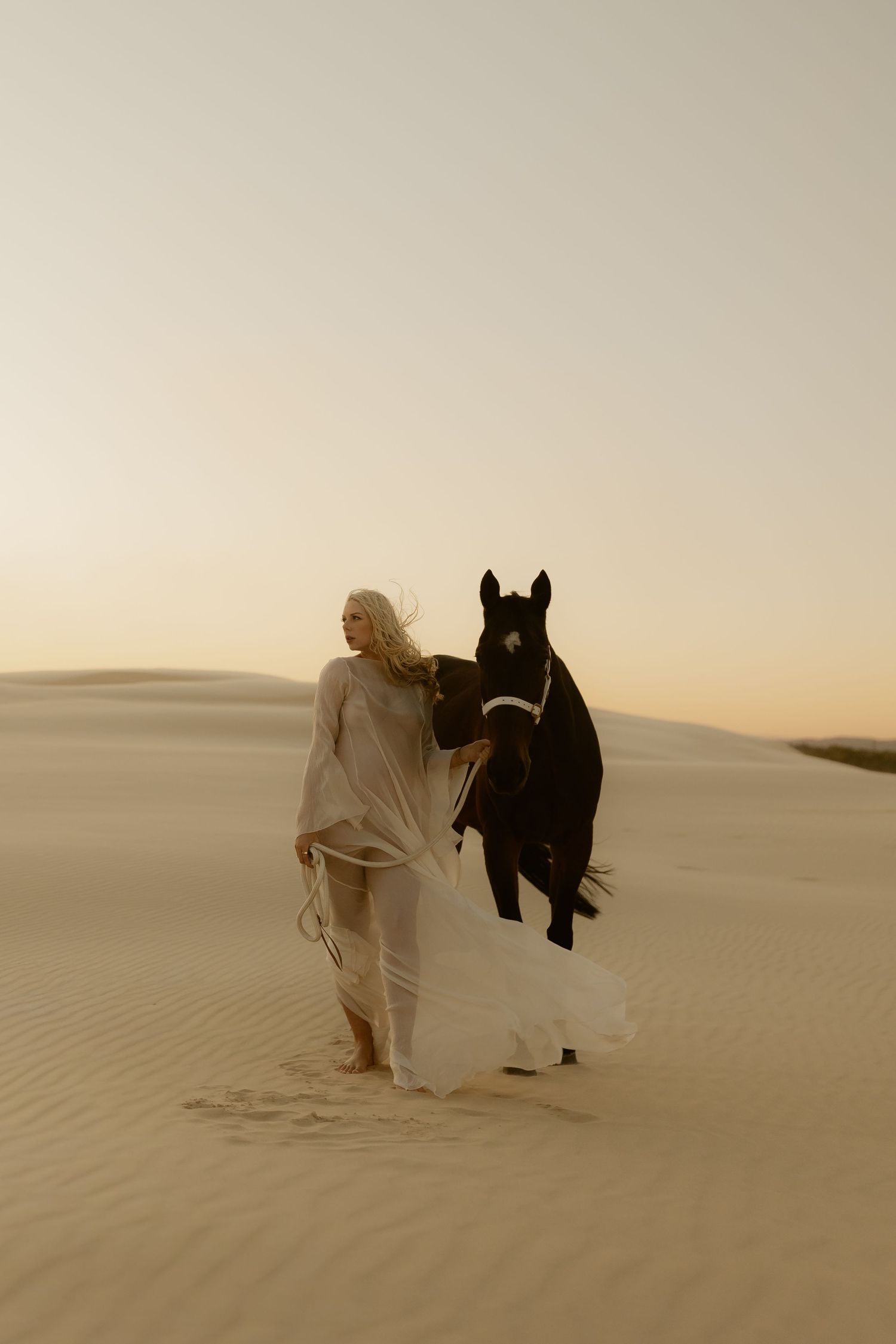 Woman in white flowing dress walks with black horse across golden sand dunes at sunset.