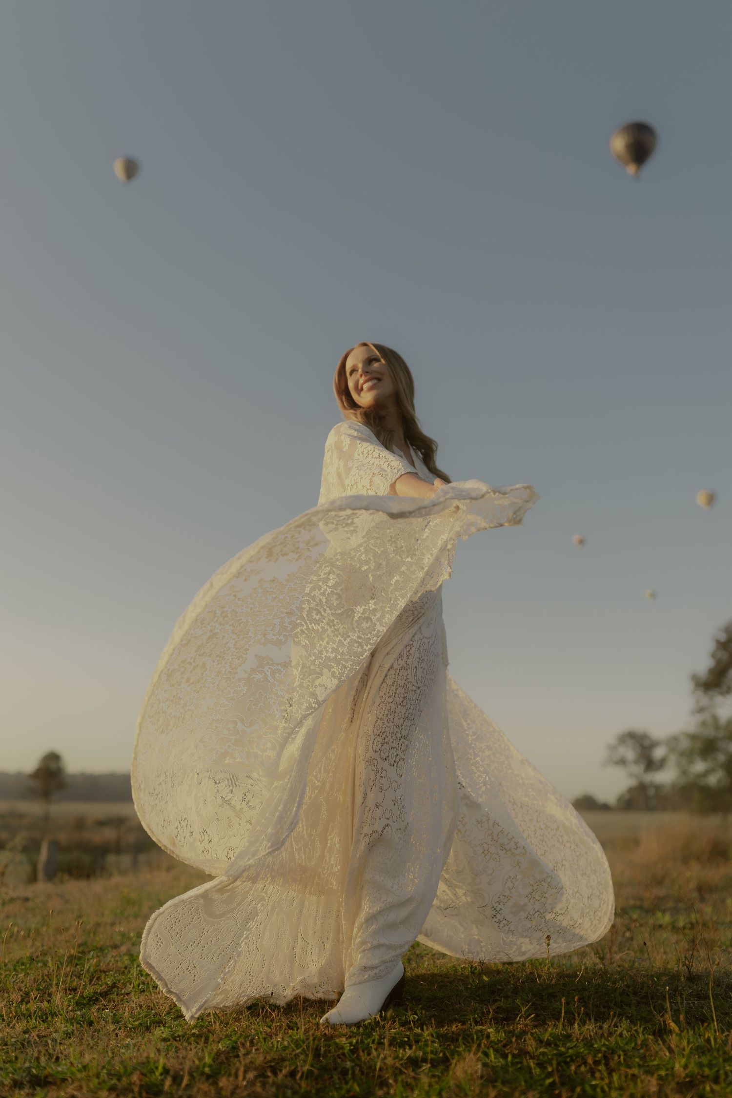 Woman in flowing white lace dress twirling in field with hot air balloons floating in sky at golden hour.