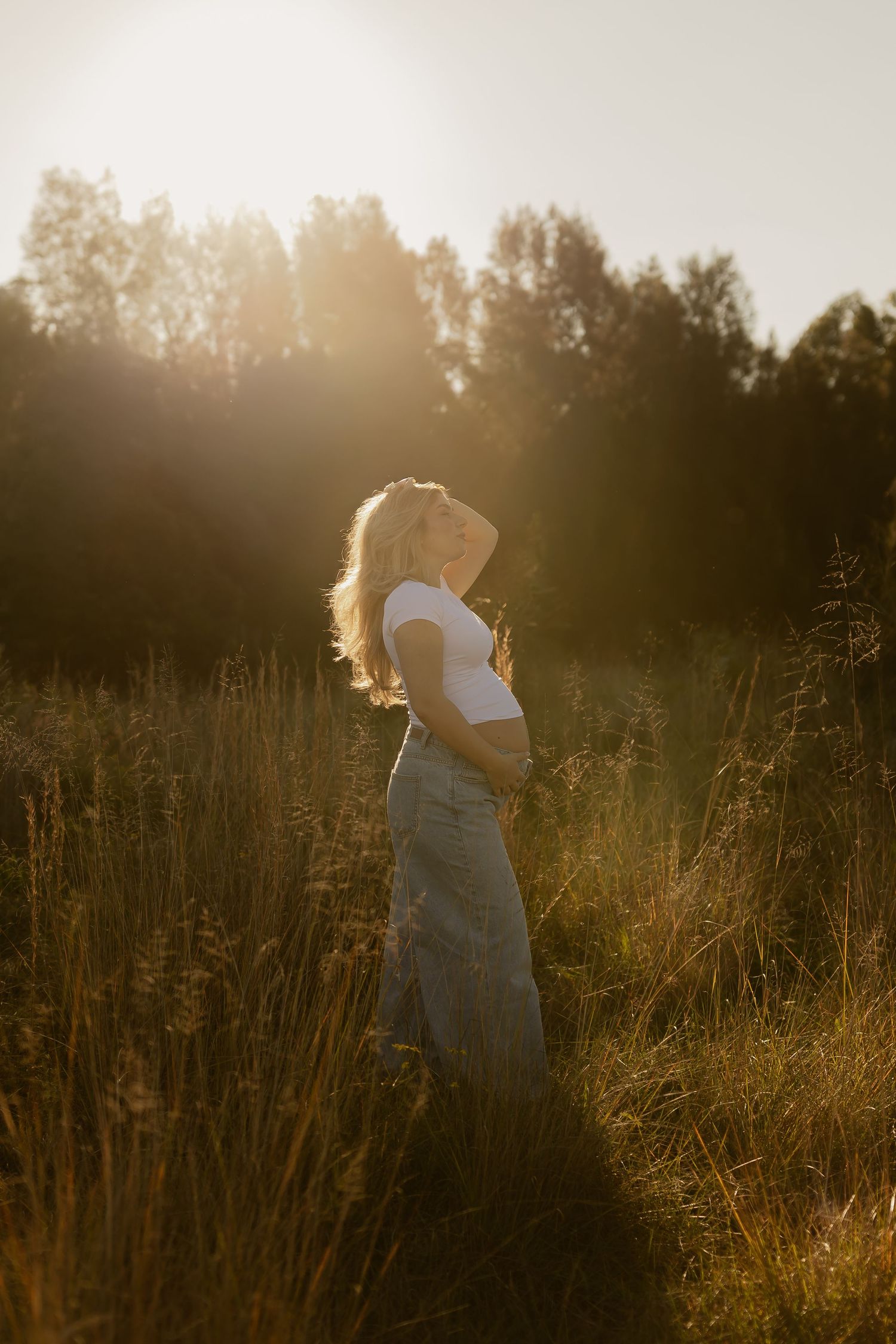 Pregnant woman in white top and jeans standing in golden sunlit field with tall grass and trees at sunset.