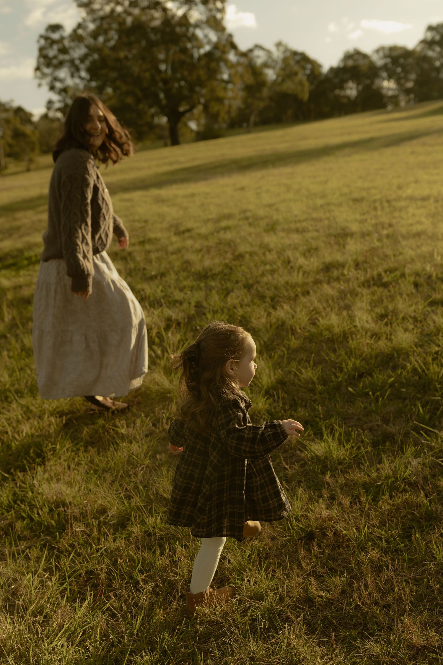 A young girl in a plaid dress runs toward a woman in a white dress on a sunlit green hillside.