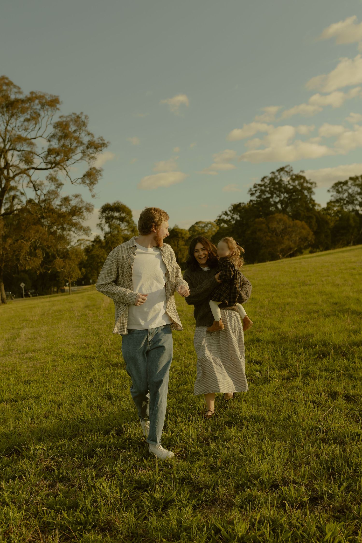 A young family walks through a sunlit green park, with a man, woman holding a toddler, and trees in the background.