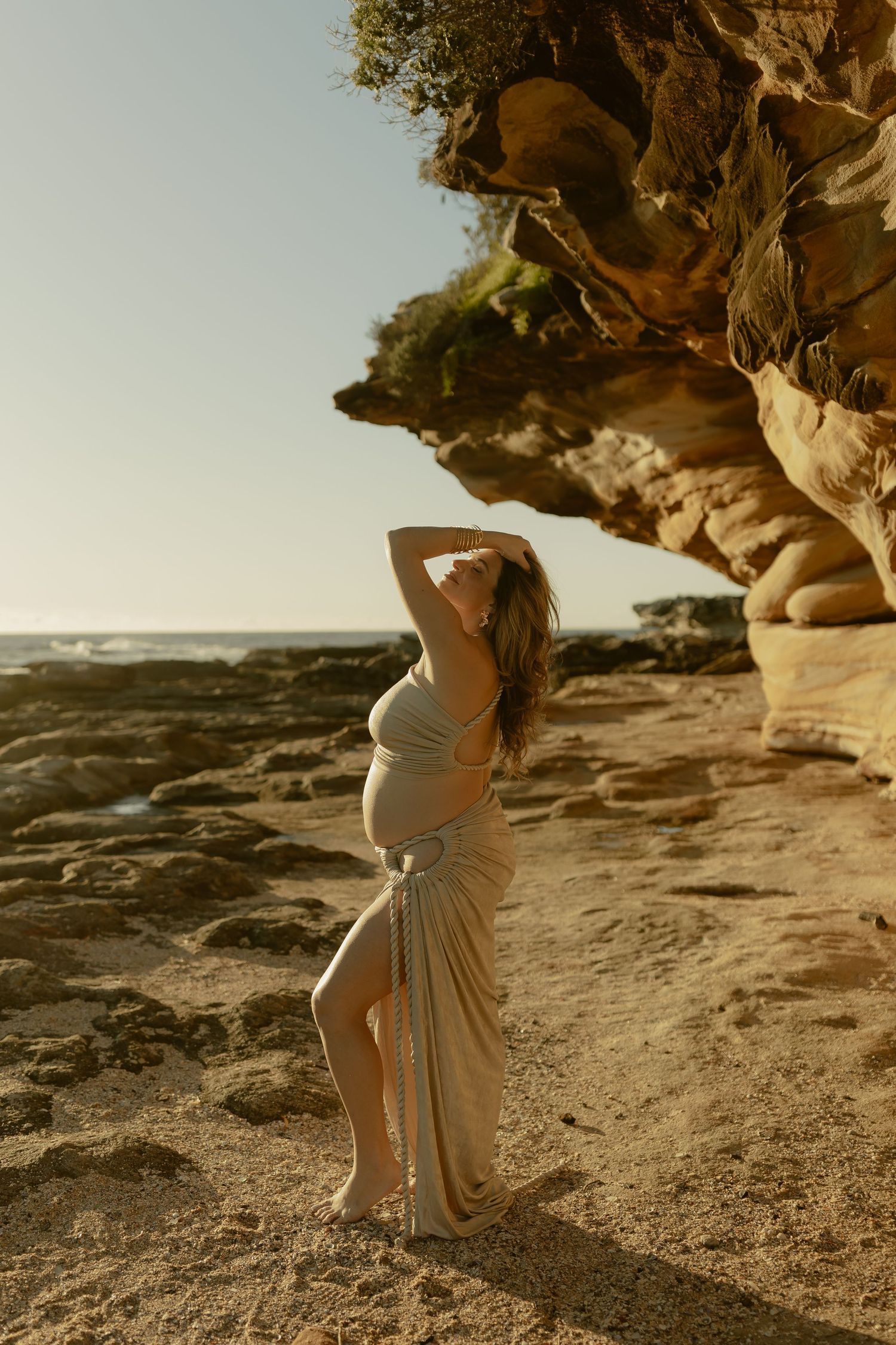 Pregnant woman in flowing beige dress poses gracefully on sandy beach near rocky cliffs at golden hour.