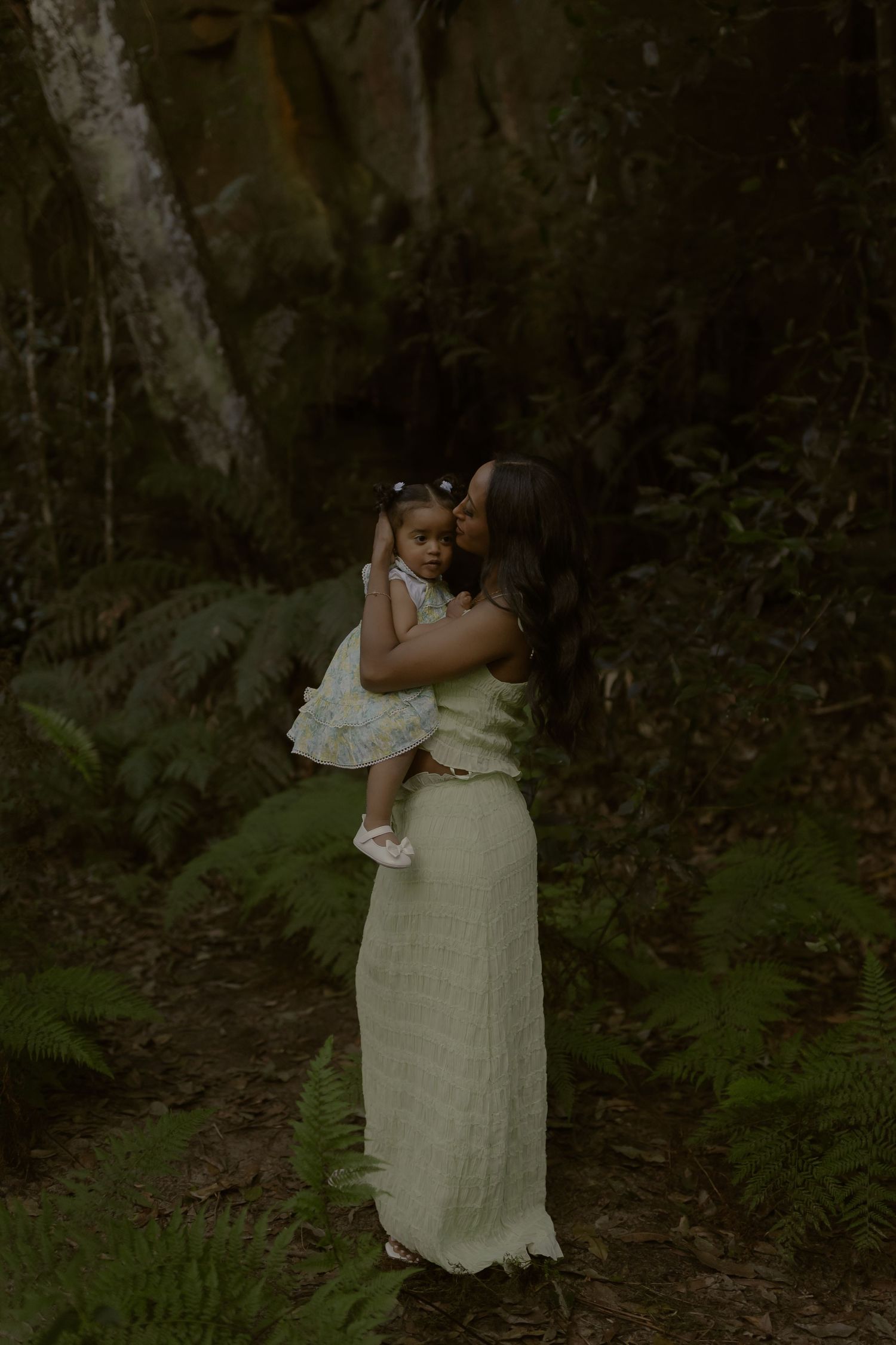 A mother in a green dress holds her toddler daughter in a lush forest setting.