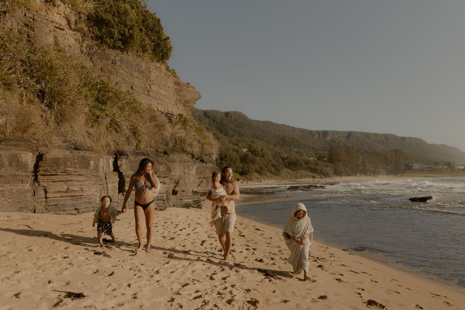 People walking along a scenic beach with rocky cliffs and coastline in the background.