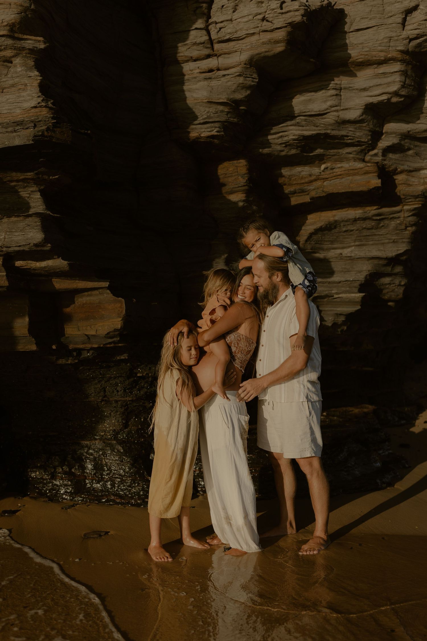 Three figures in light dresses embrace joyfully against a dramatic rocky cave backdrop.