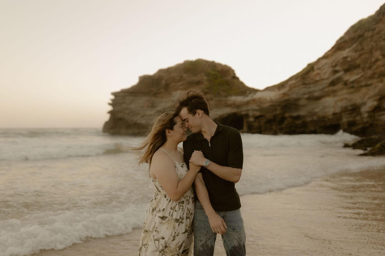 Romantic beach photoshoot with rocky cliffs and ocean waves in the background at sunset.