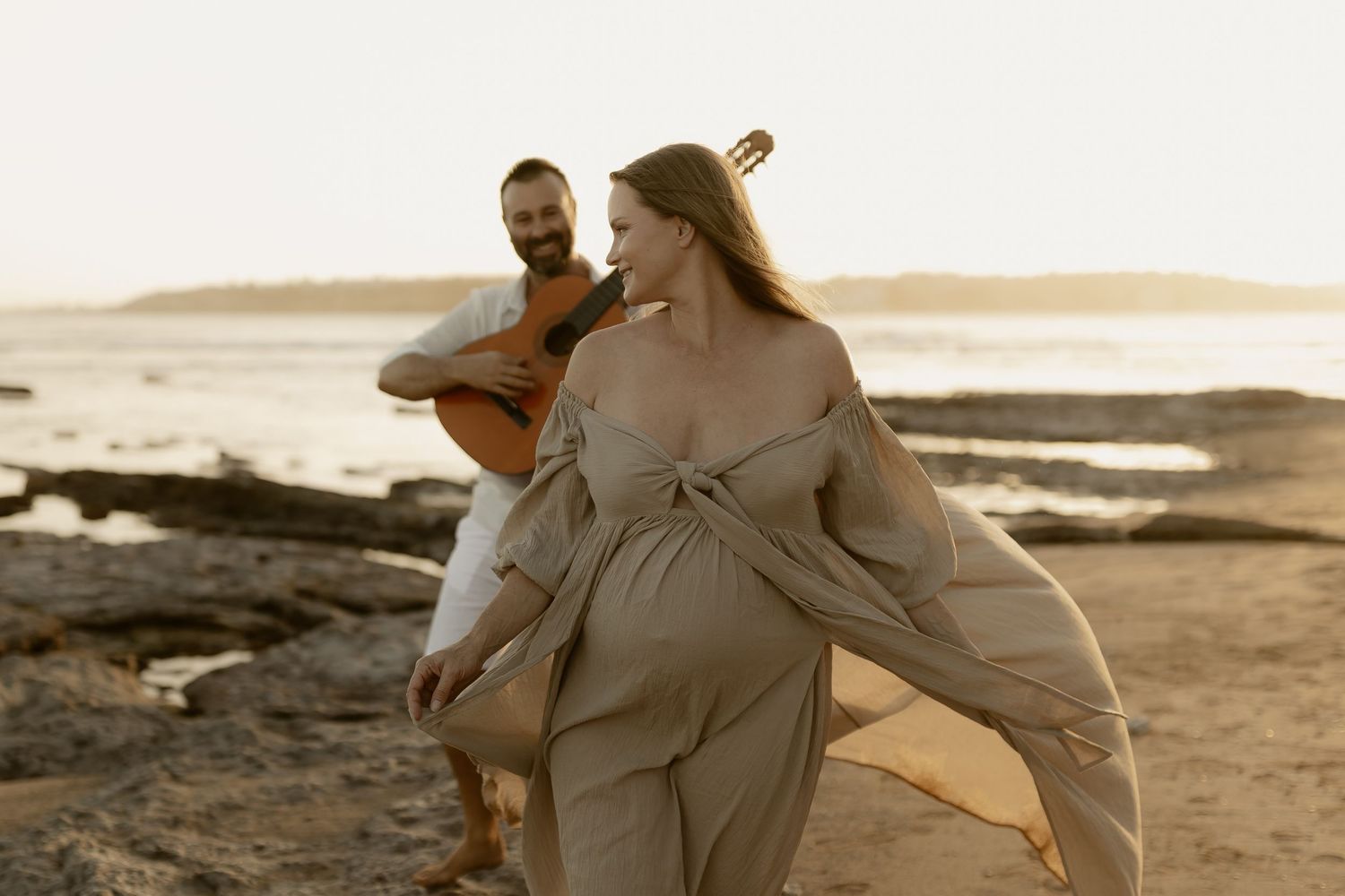 A couple embraces on a sandy beach during golden hour as waves roll in along the shoreline.