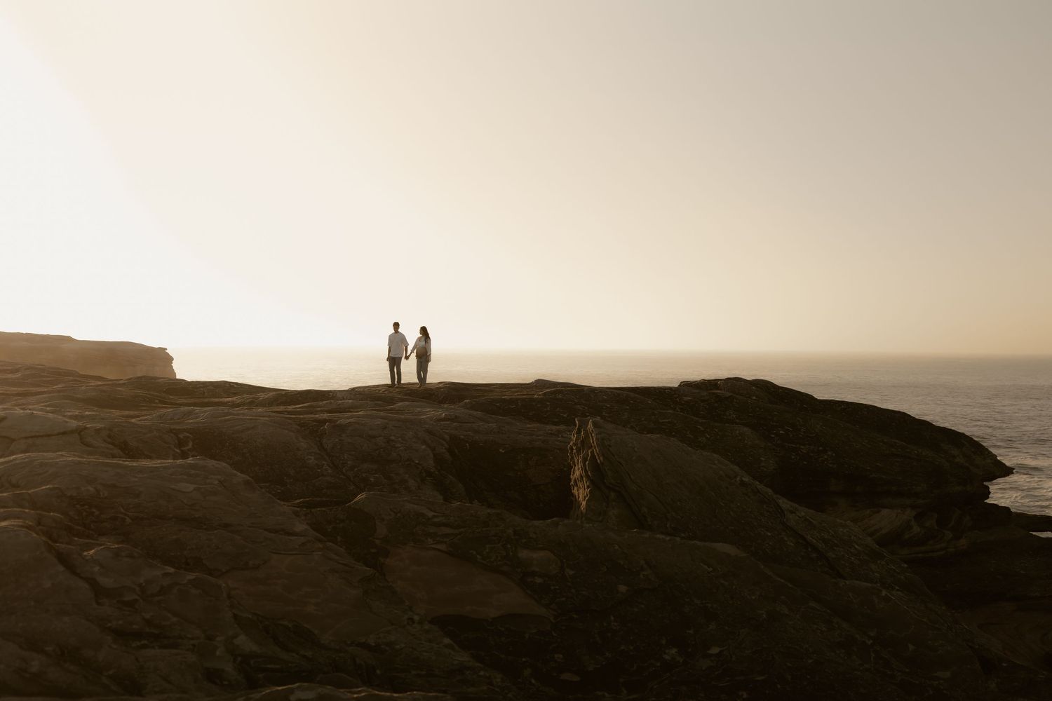 Two silhouettes stand atop a dramatic coastal cliff against a misty sunset backdrop.