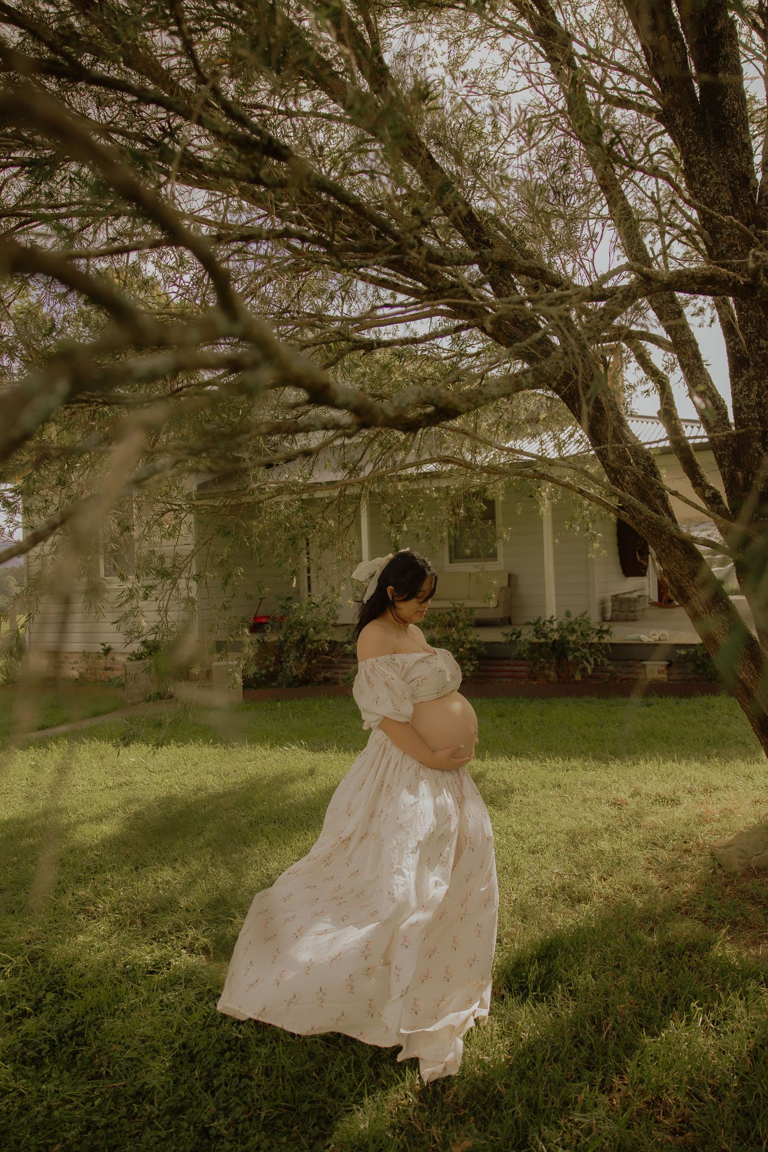 Pregnant woman in flowing white off-shoulder gown standing among trees in garden.