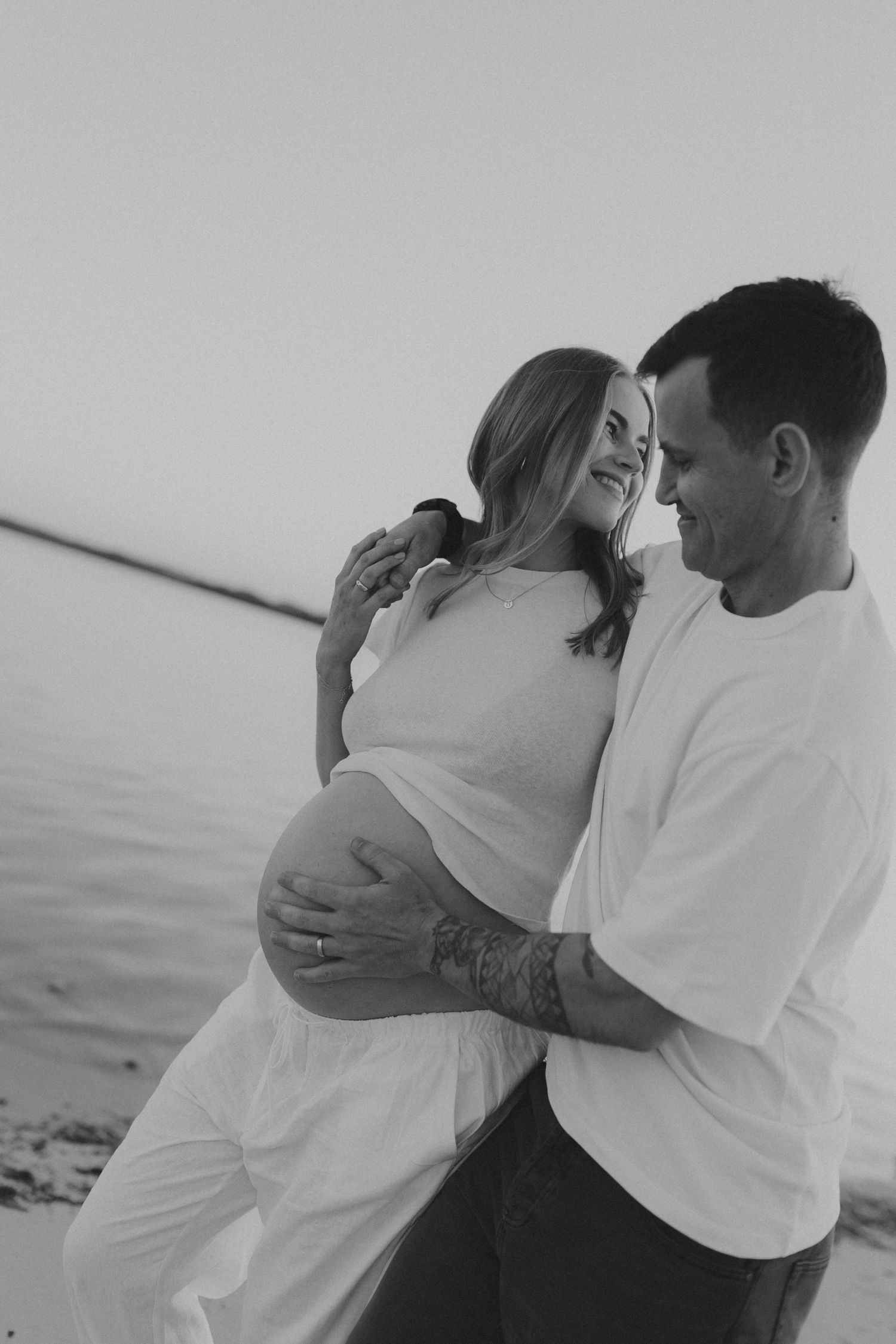 Black and white photo of a couple embracing on a beach, man holding pregnant womans belly near the water.