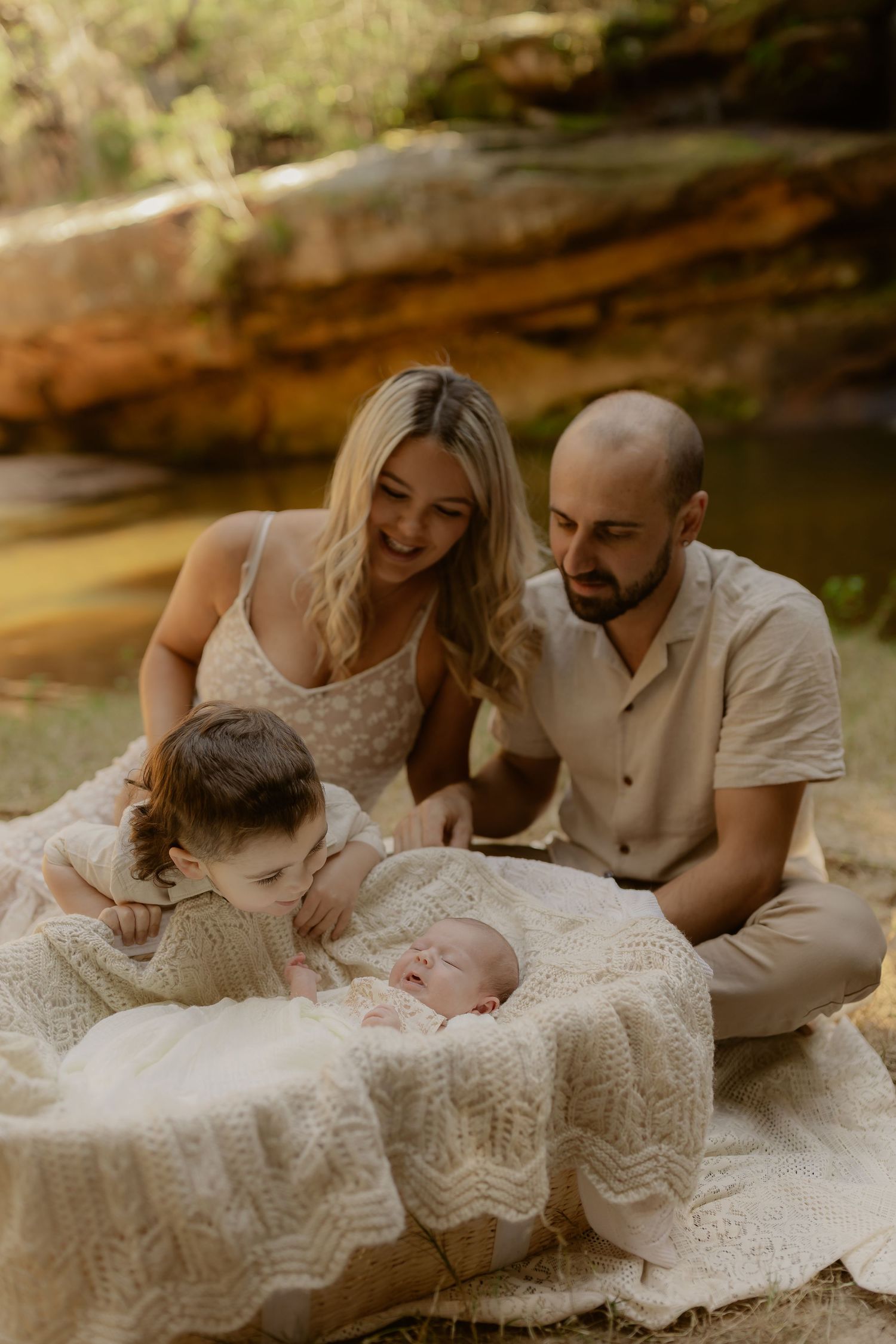 A young family sits on a blanket outdoors, parents smiling at their newborn baby while a toddler looks on.