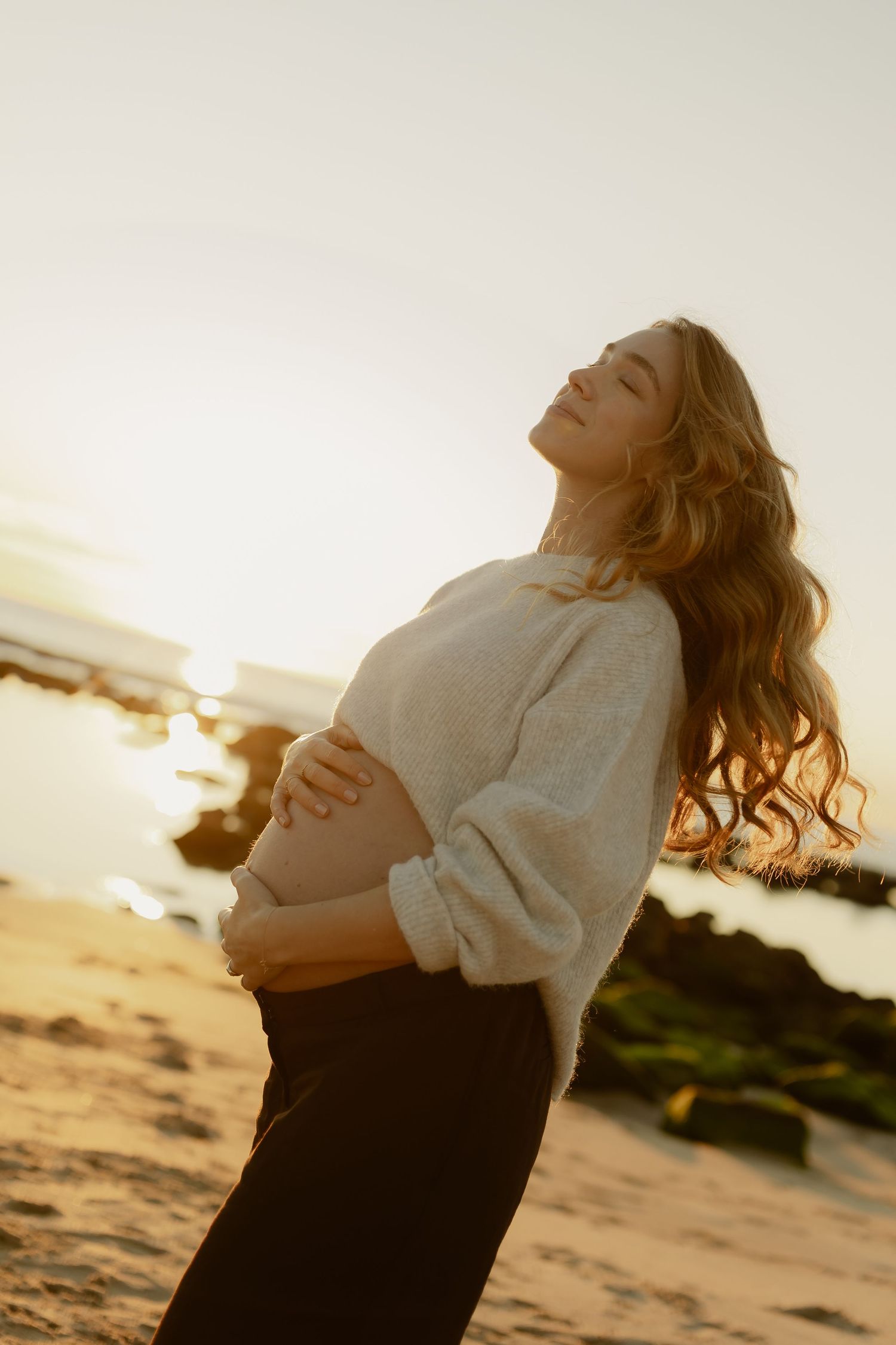 Pregnant woman in cream sweater cradles baby bump on beach, eyes closed, bathed in golden sunset light.