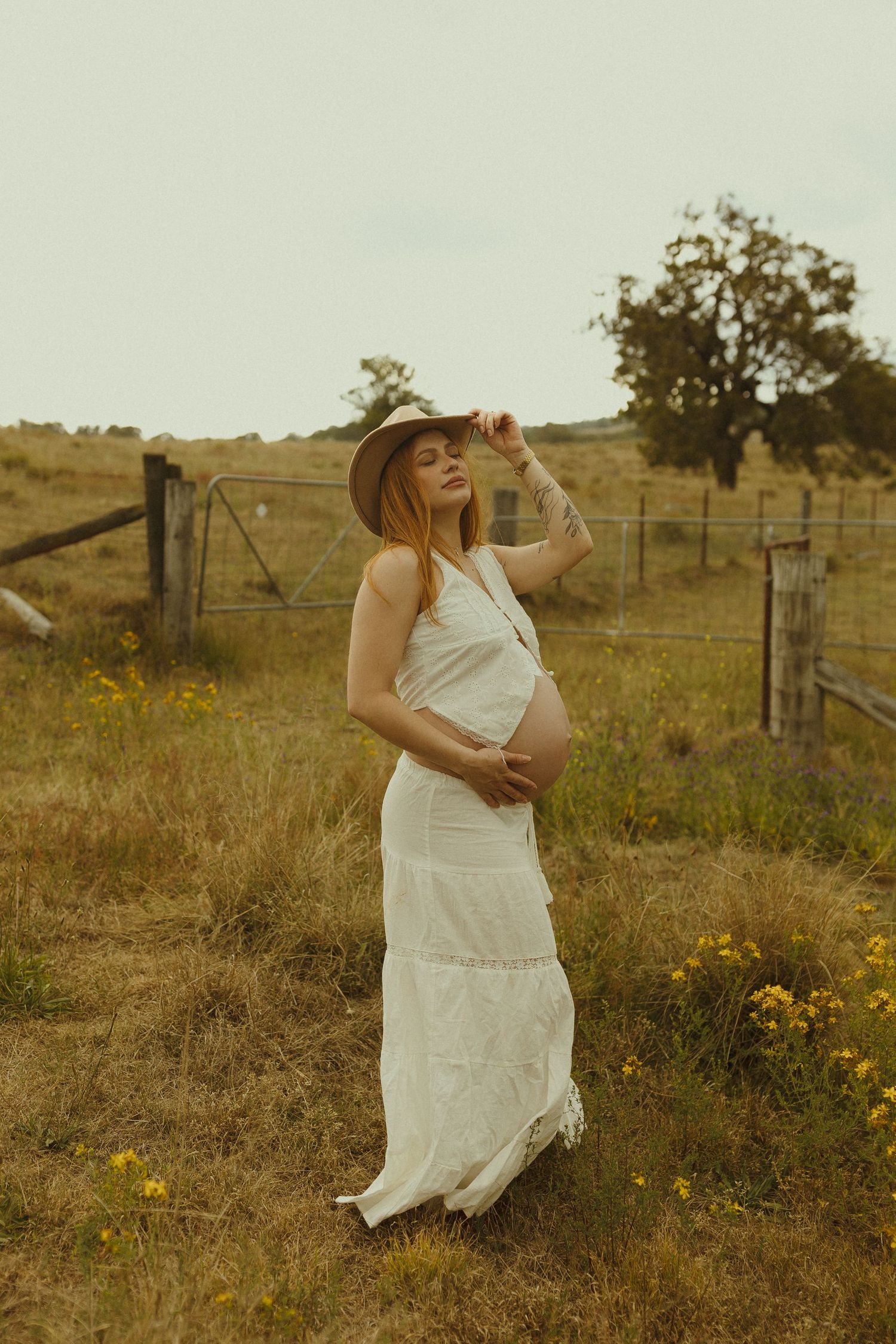 A countryside maternity photo featuring a white dress and cowboy hat against rolling hills.