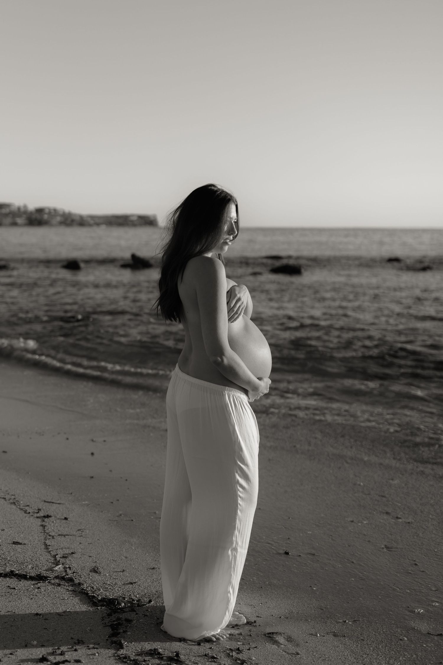 A black and white portrait of someone in white clothing standing thoughtfully by the ocean.