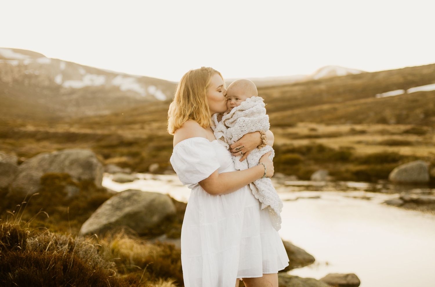 A family wearing white outfits embraces during a golden sunset photoshoot in a scenic mountain landscape.