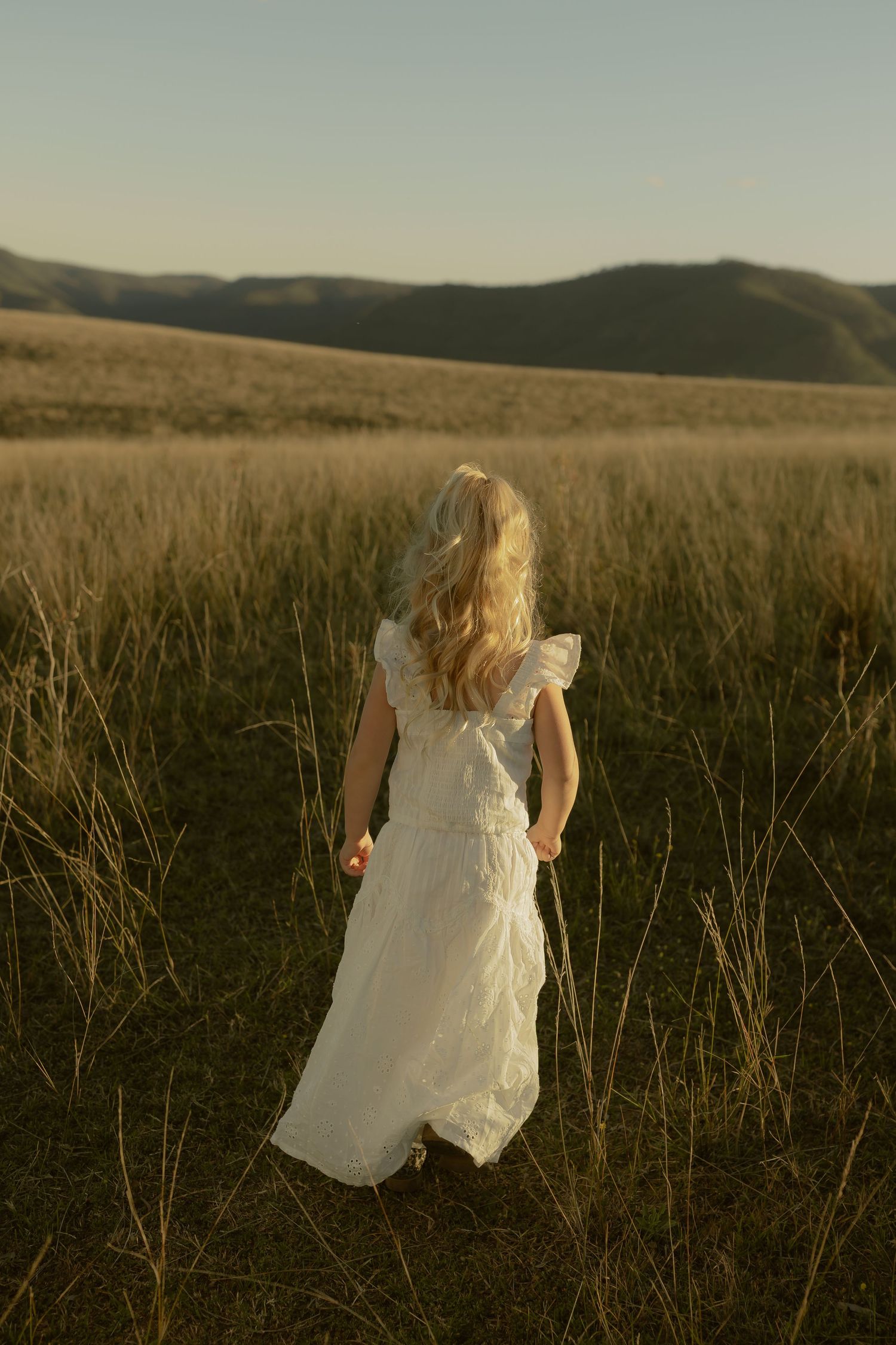 Young girl in white dress walking through golden field with rolling hills in background.