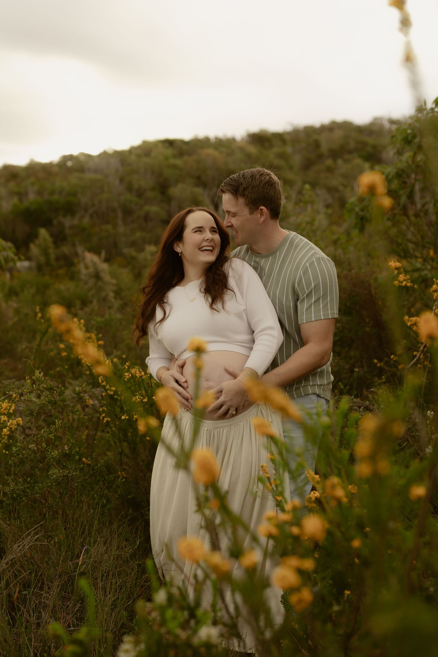 Couple sharing a joyful maternity moment outdoors, surrounded by yellow wildflowers and lush greenery.