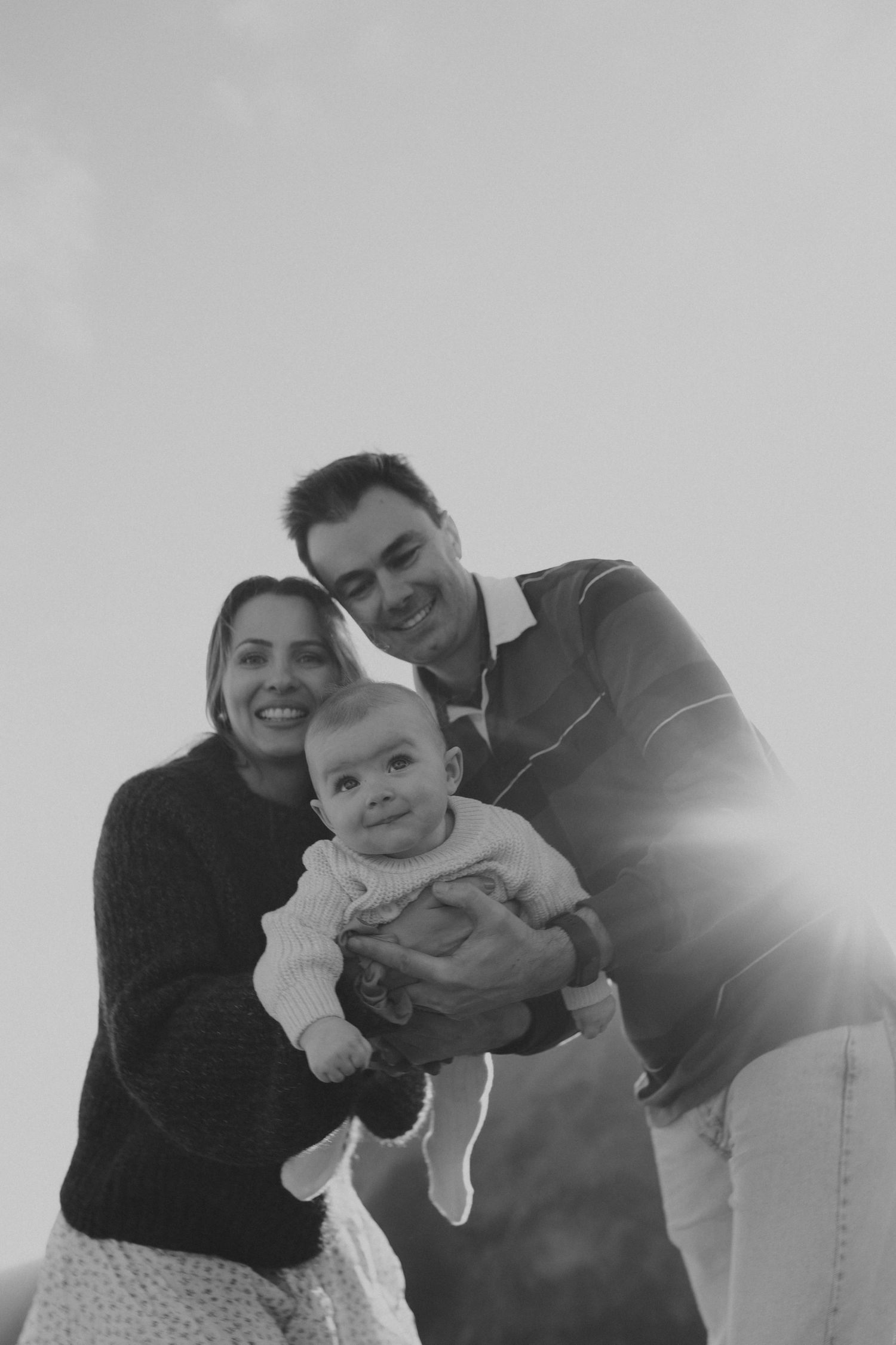 Black and white family portrait of smiling parents holding their baby with sunlight behind them.