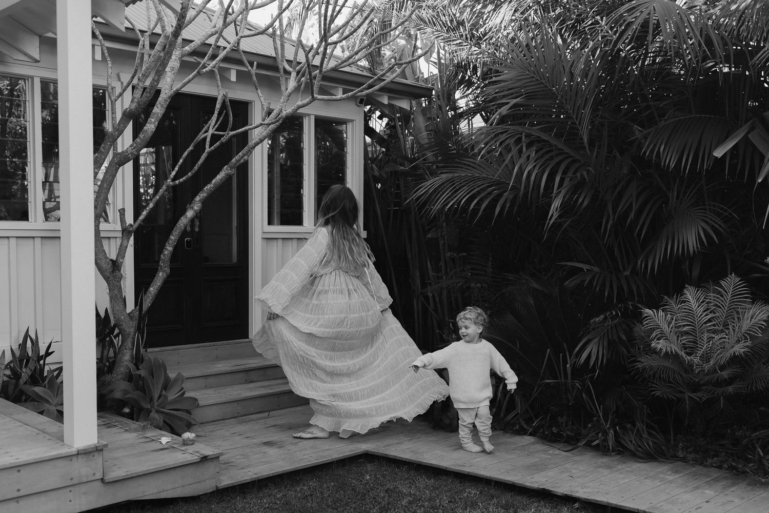 Black and white photo of child running on porch near tropical plants.