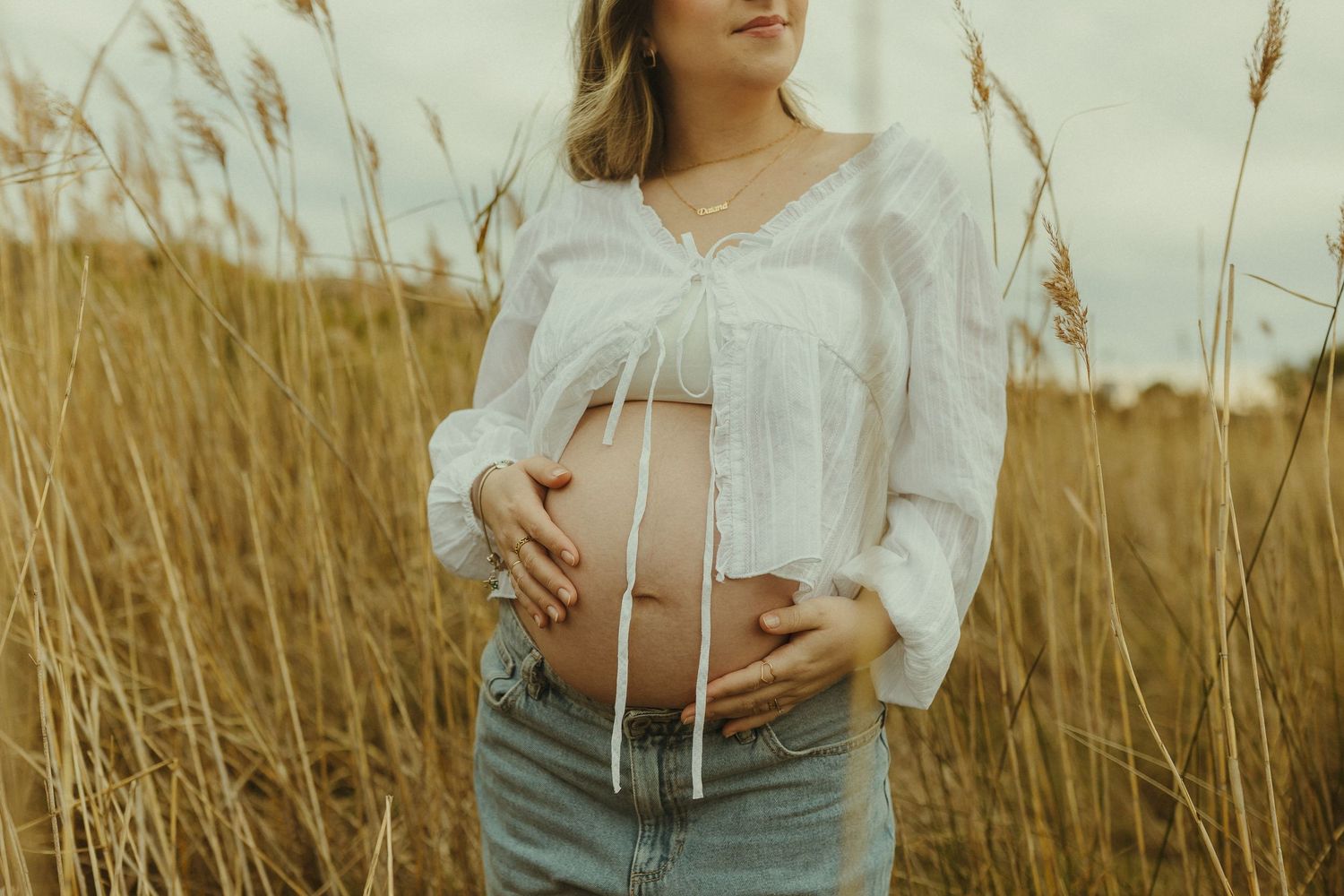 Person in a white flowing dress poses in a golden wheat field at sunset, showcasing maternity style.