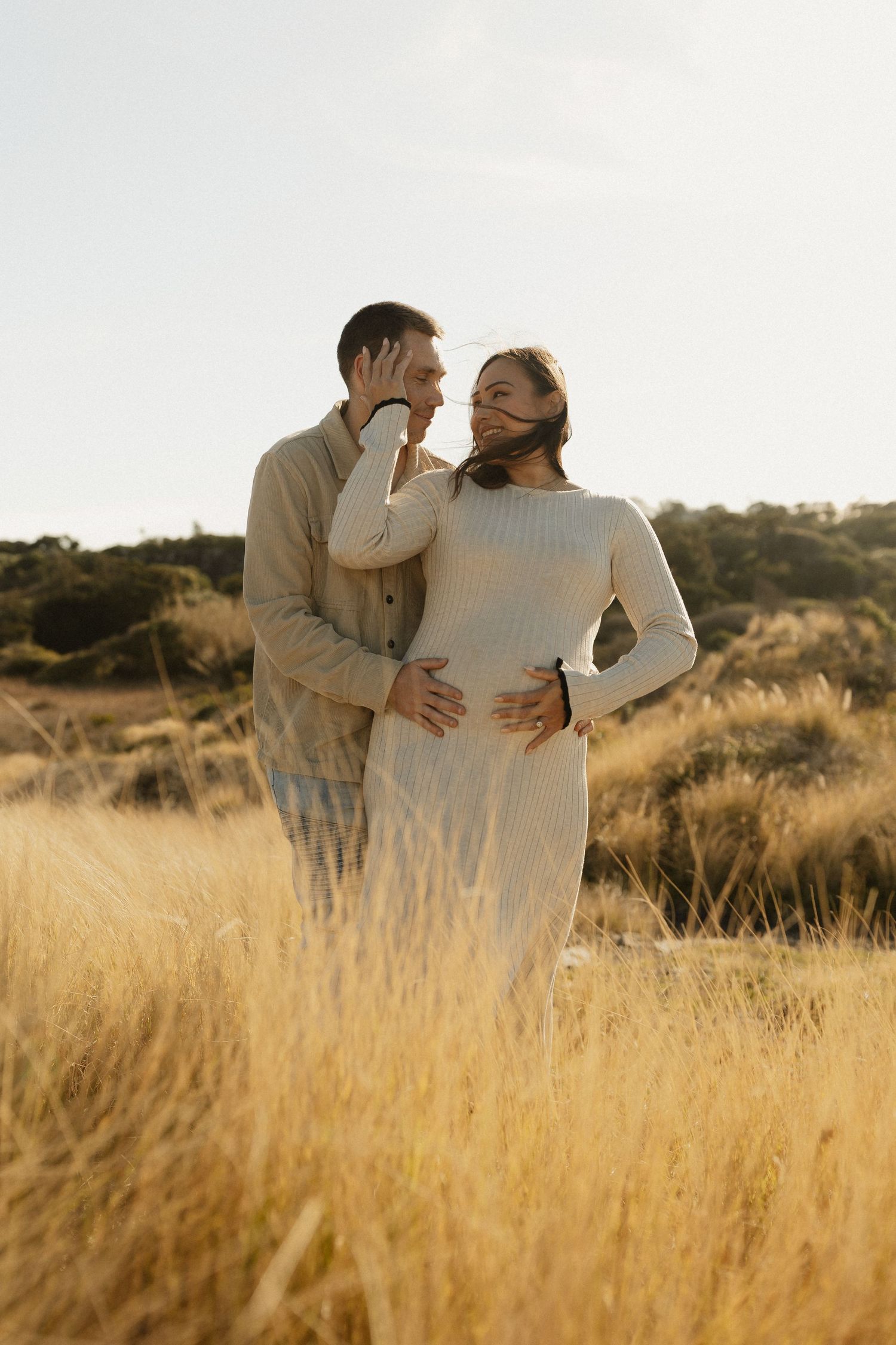 A loving couple embraces in a golden field during sunset wearing neutral colored clothing.