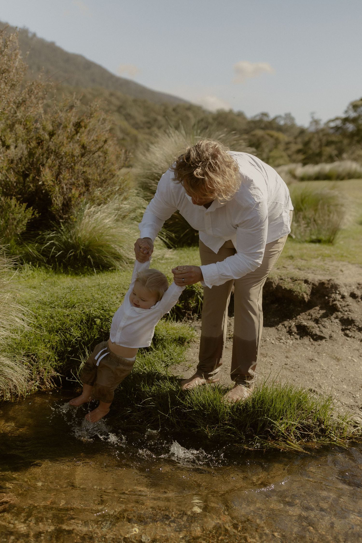 Two people in white shirts explore a stream in a natural setting on a sunny day.