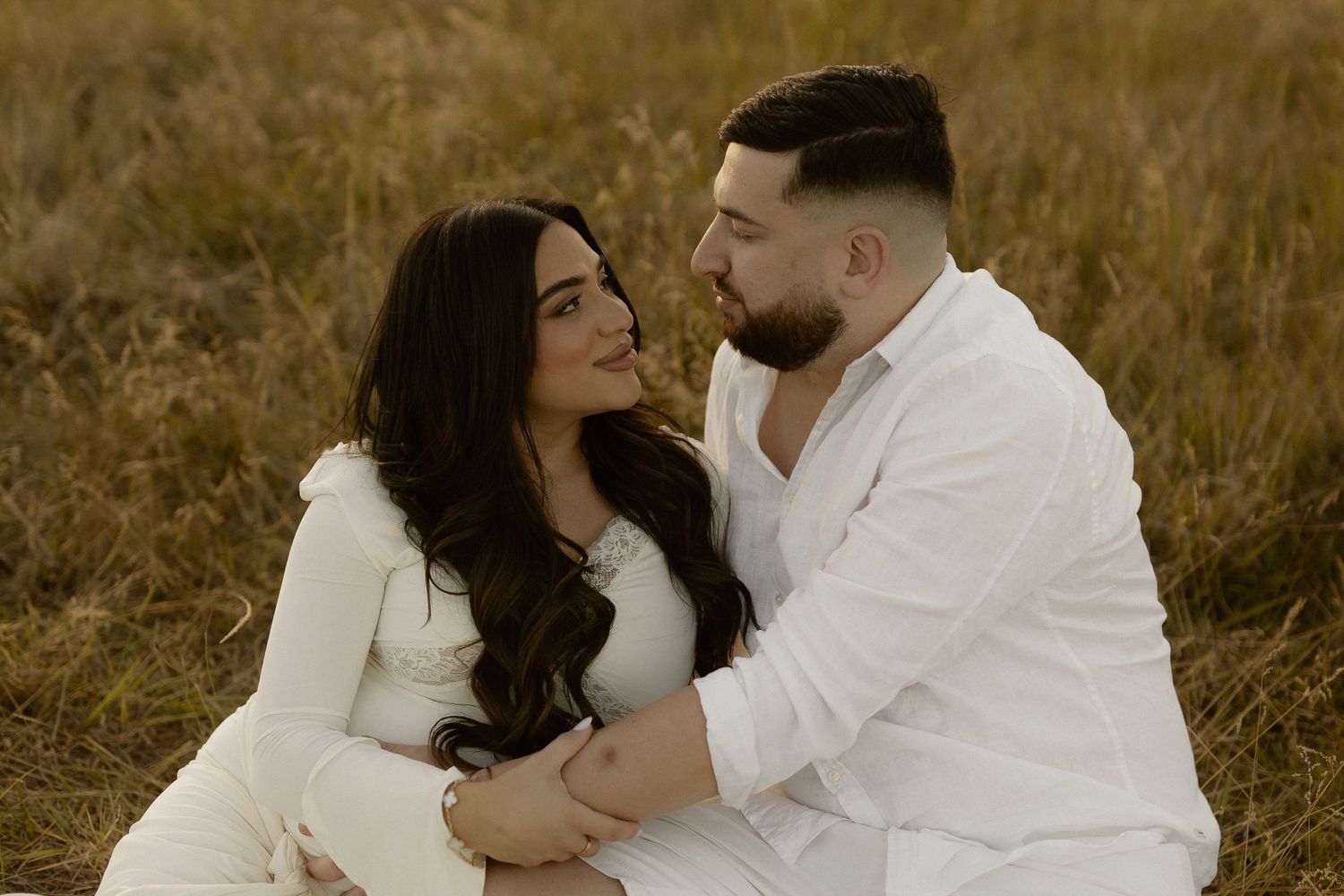 A couple in white outfits sits together intimately in a golden field during sunset.
