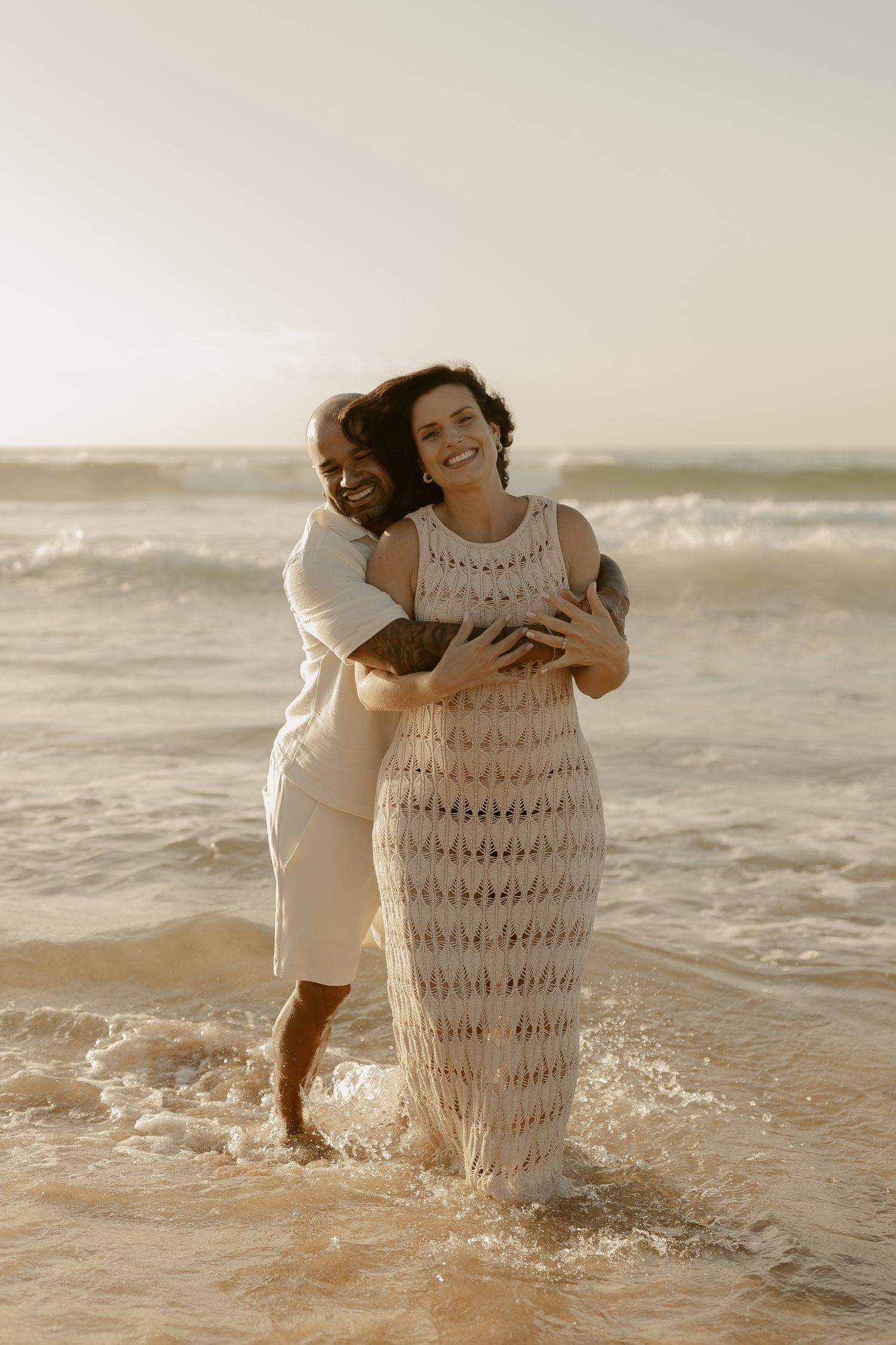 Intimate beach portrait of couple embracing in shallow ocean waves at sunset.