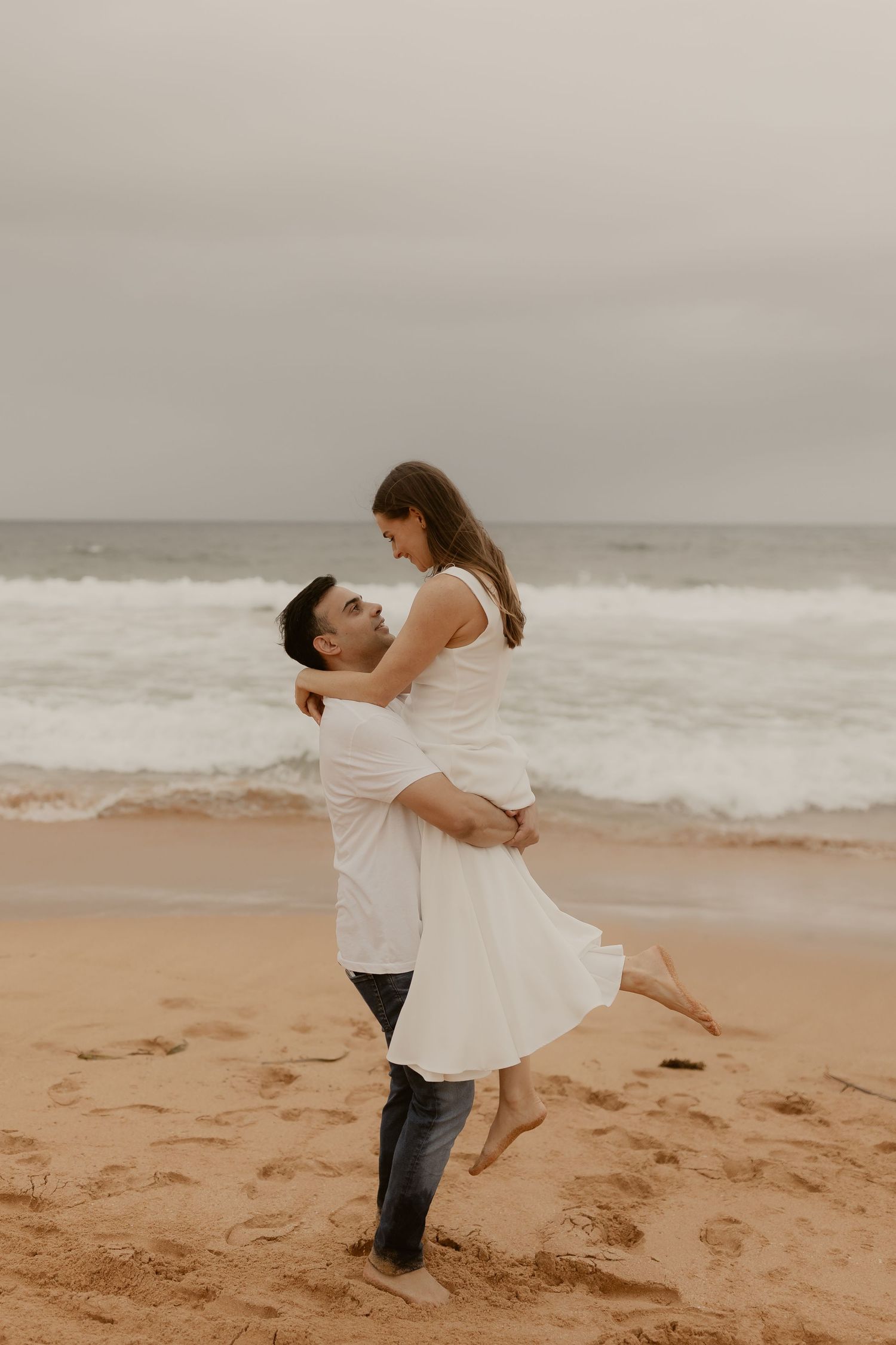 A couple shares a romantic dance moment on a misty beach with waves rolling in.
