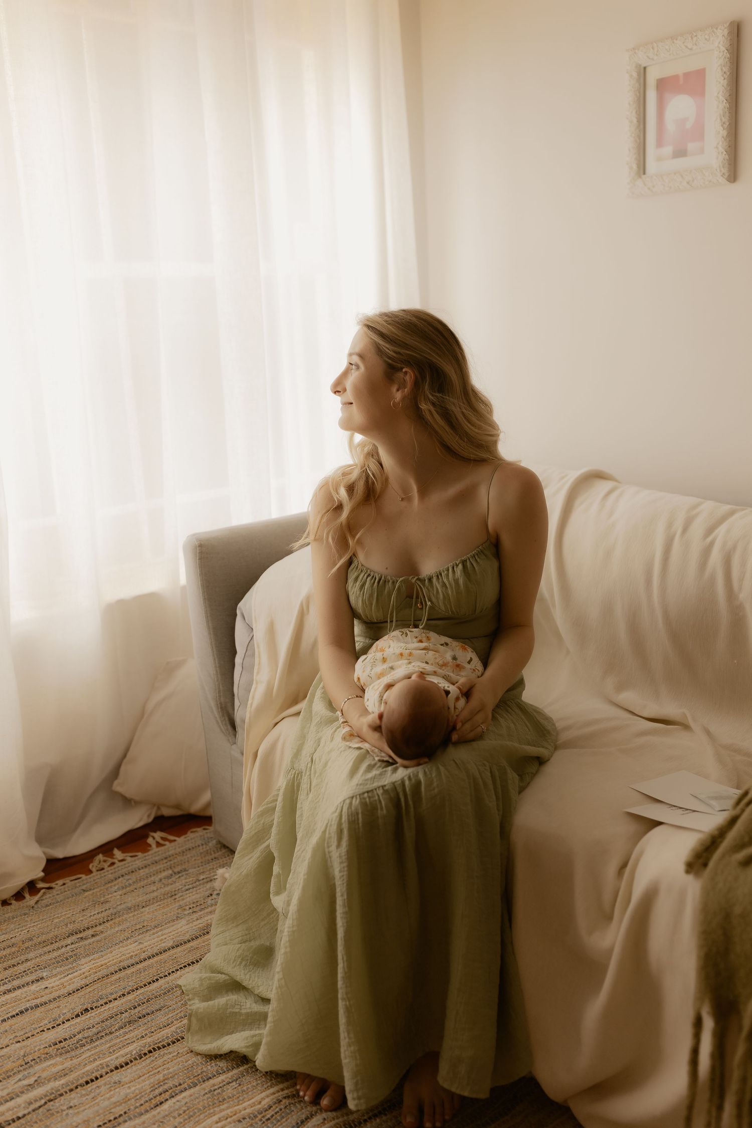 Person in sage green dress sitting on white couch in sunlit room with baby.
