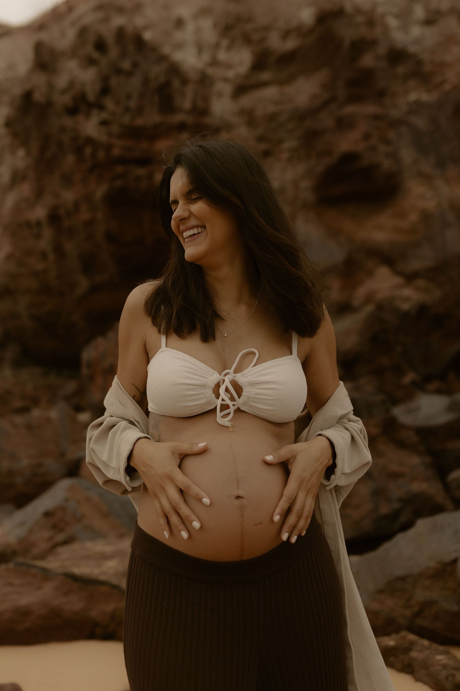 Artistic maternity photo at sunset with expecting mother in white crop top and flowing dress against rocky backdrop.