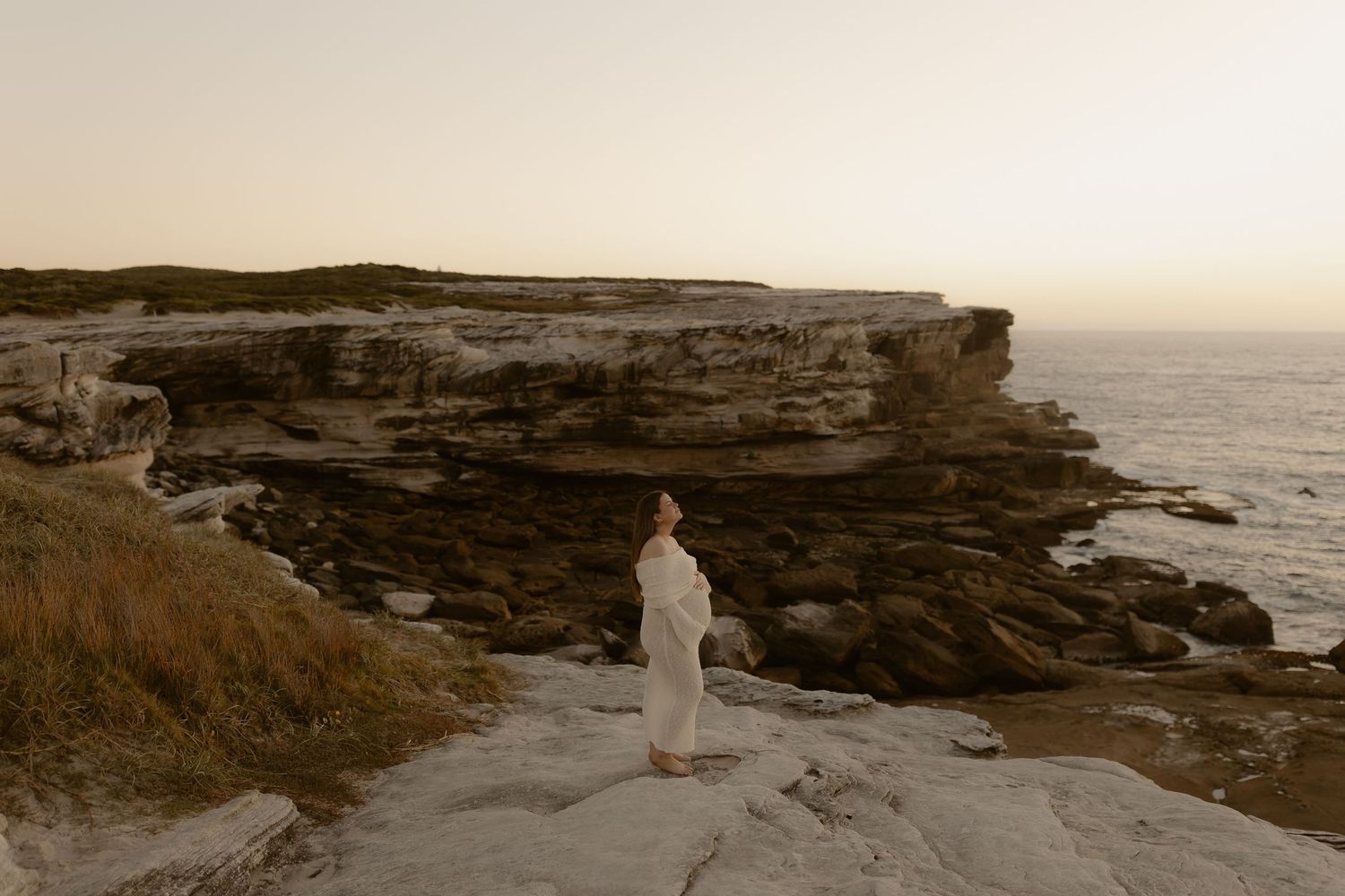 A figure in white stands on rocky coastal cliffs at sunset overlooking the dramatic ocean landscape.