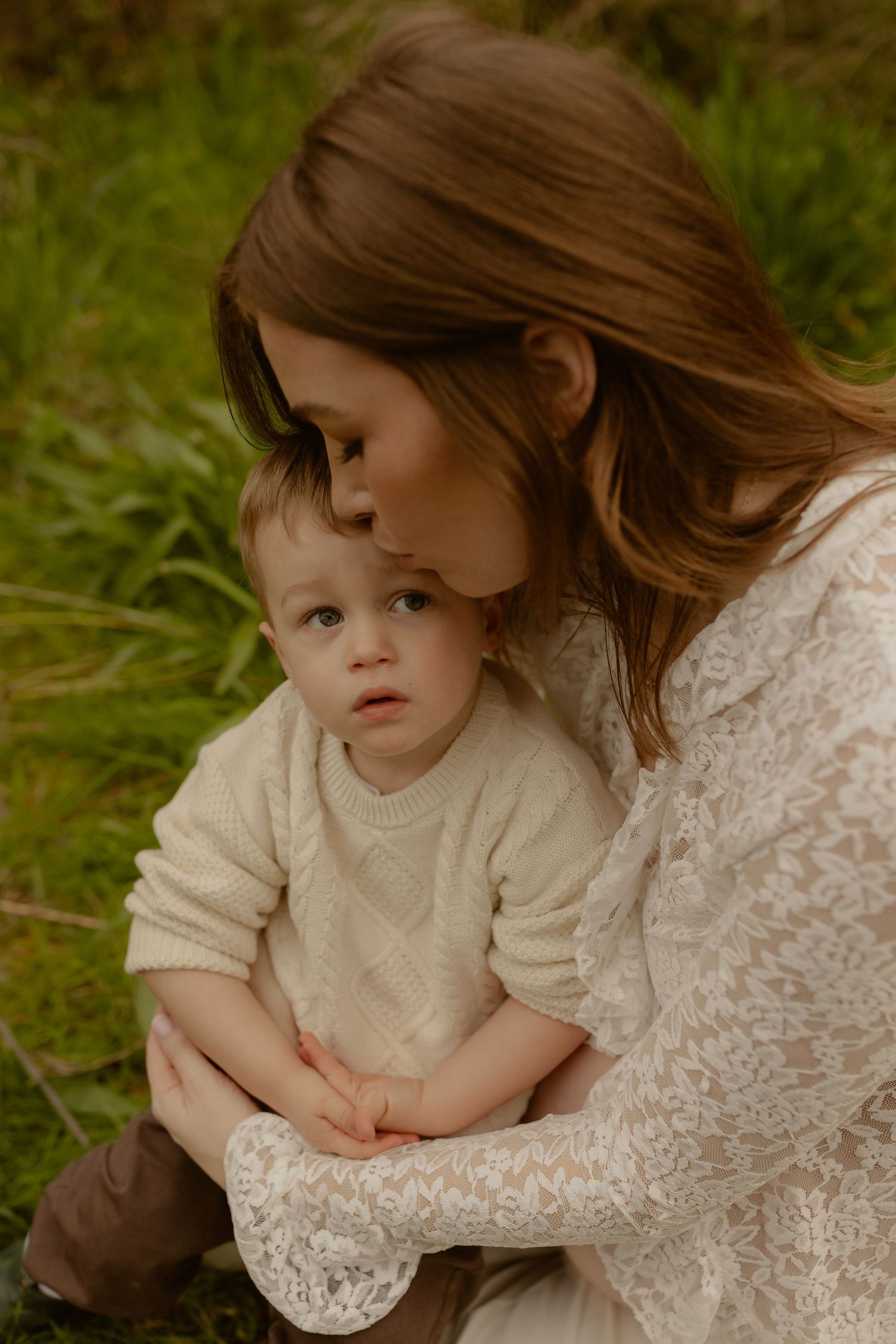 A mother gently kisses her toddler's head while holding the child close in a lush green field.