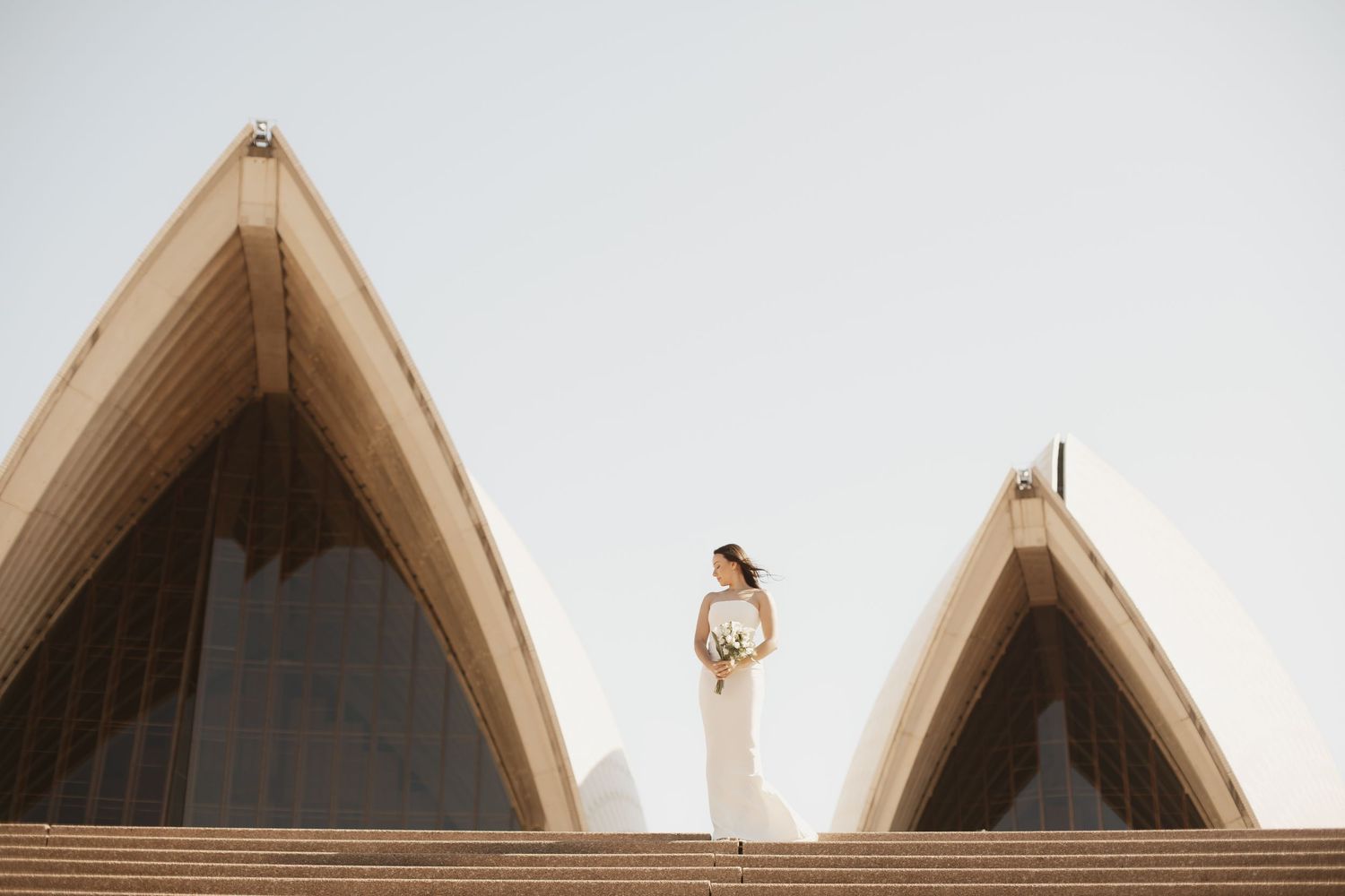A figure in a white dress stands between the iconic pointed arches of the Sydney Opera House at sunset.