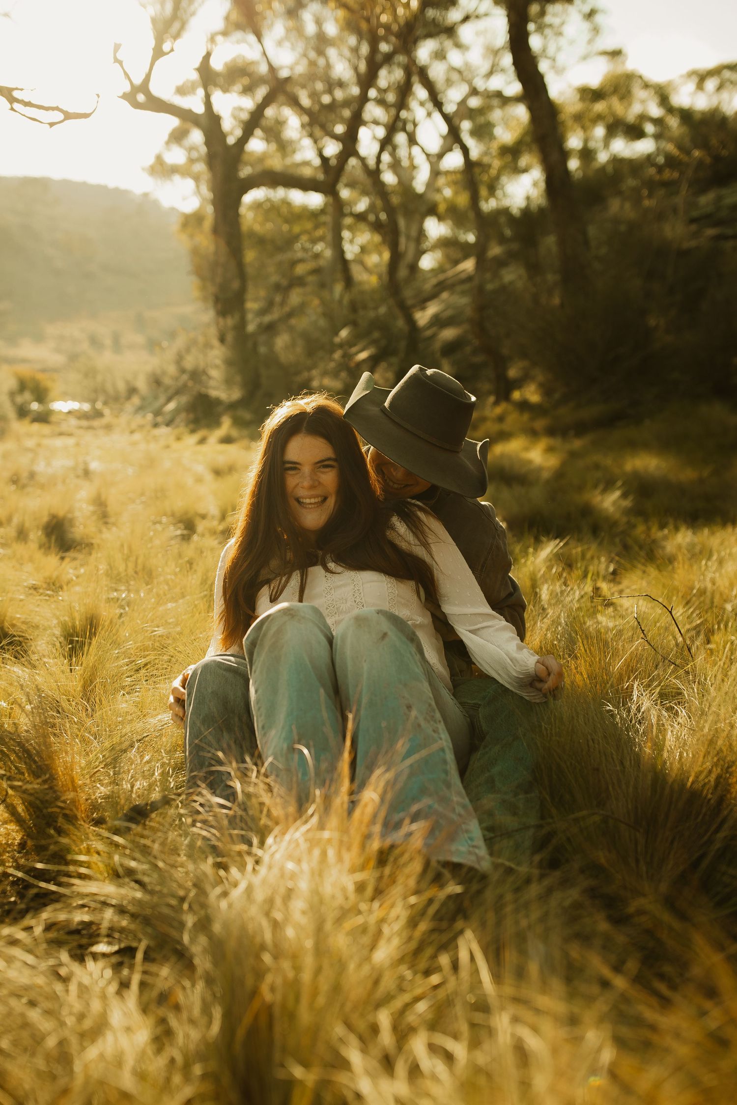 A romantic couple sits in golden grass at sunset with trees silhouetted in the background.